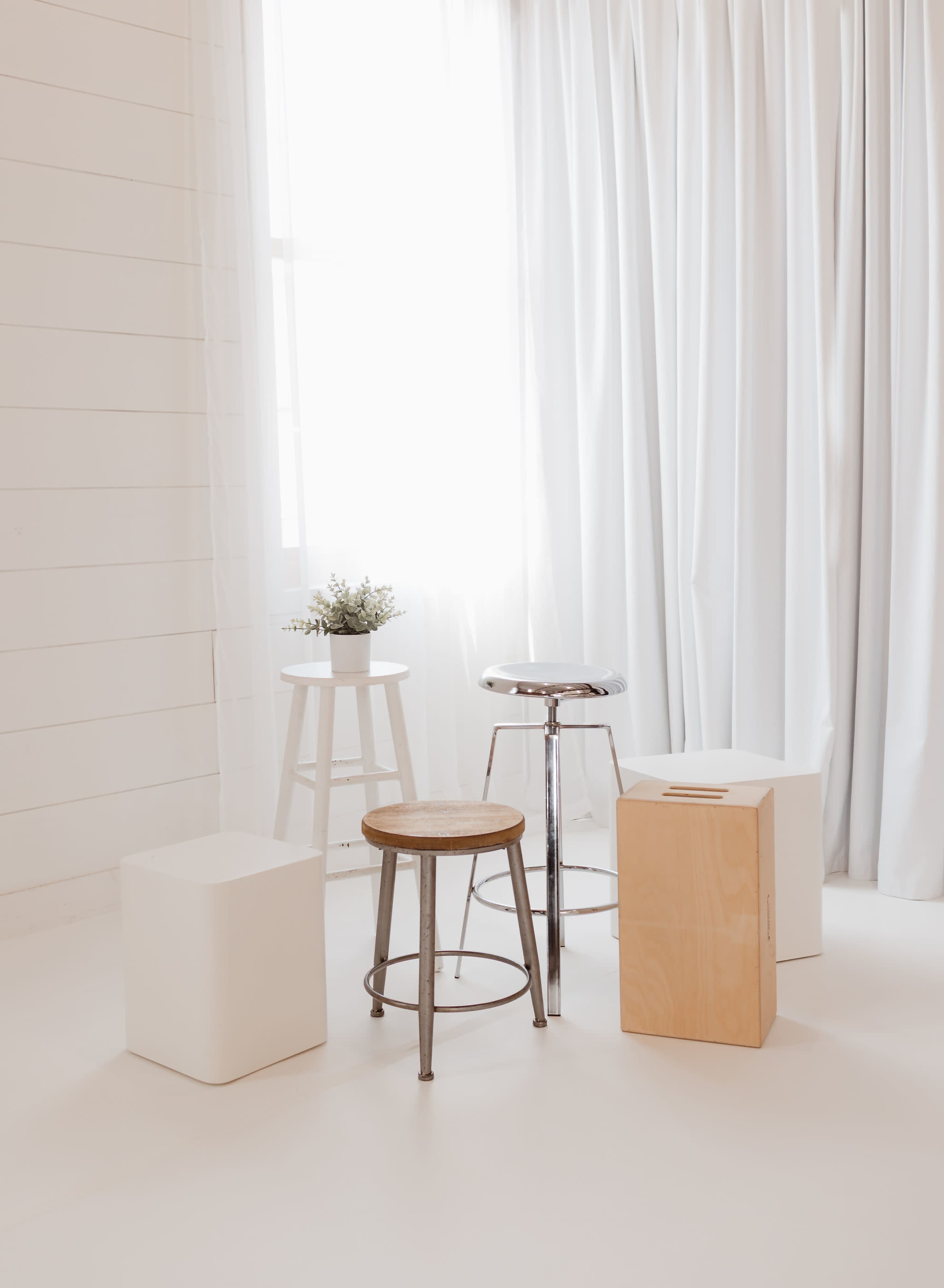 The image features a minimalist scene with four stools of varying heights positioned near a window, alongside a small potted plant on one of the stools.