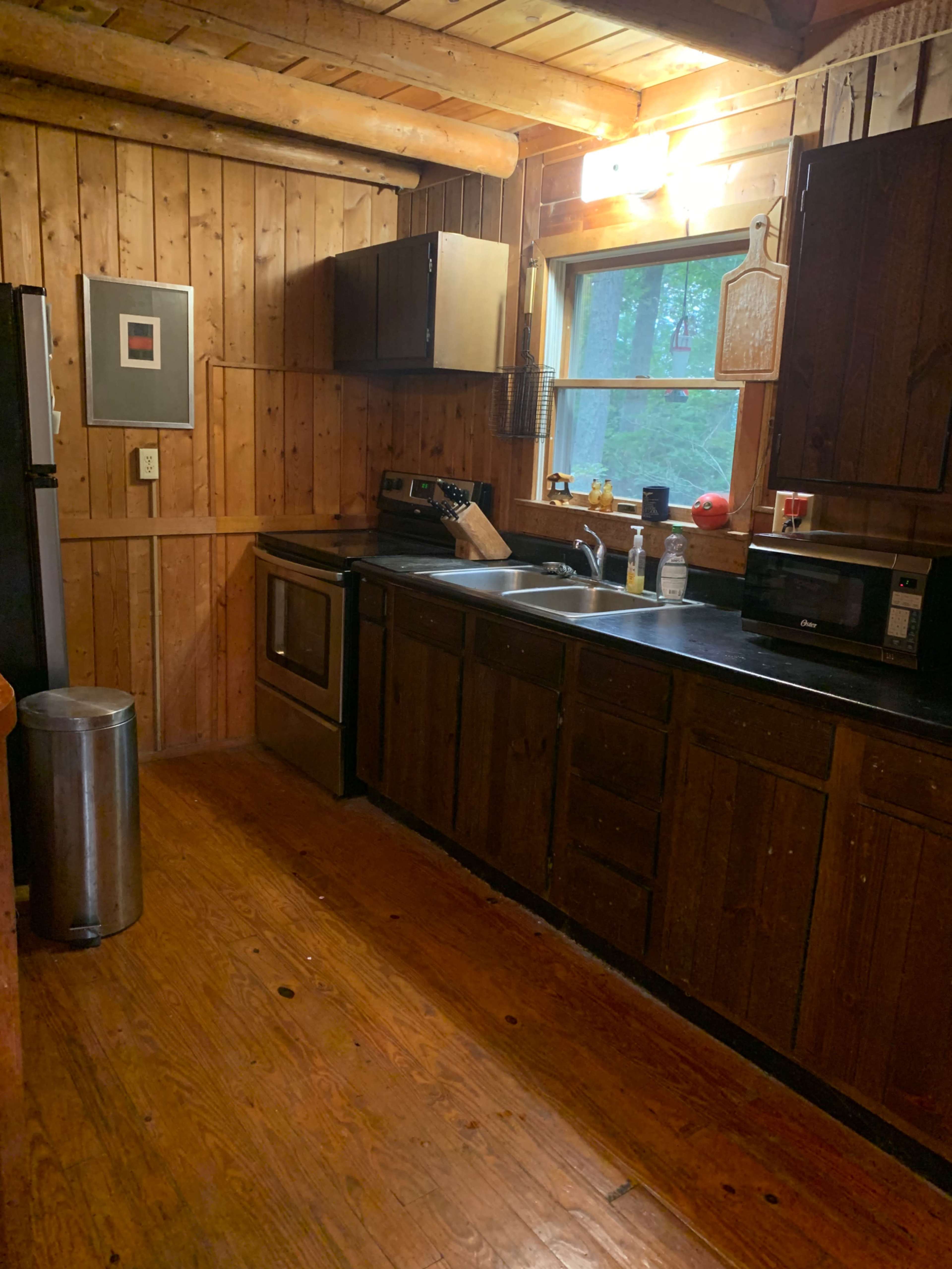A rustic kitchen with wooden walls, a stainless steel stove, and a window overlooking greenery.