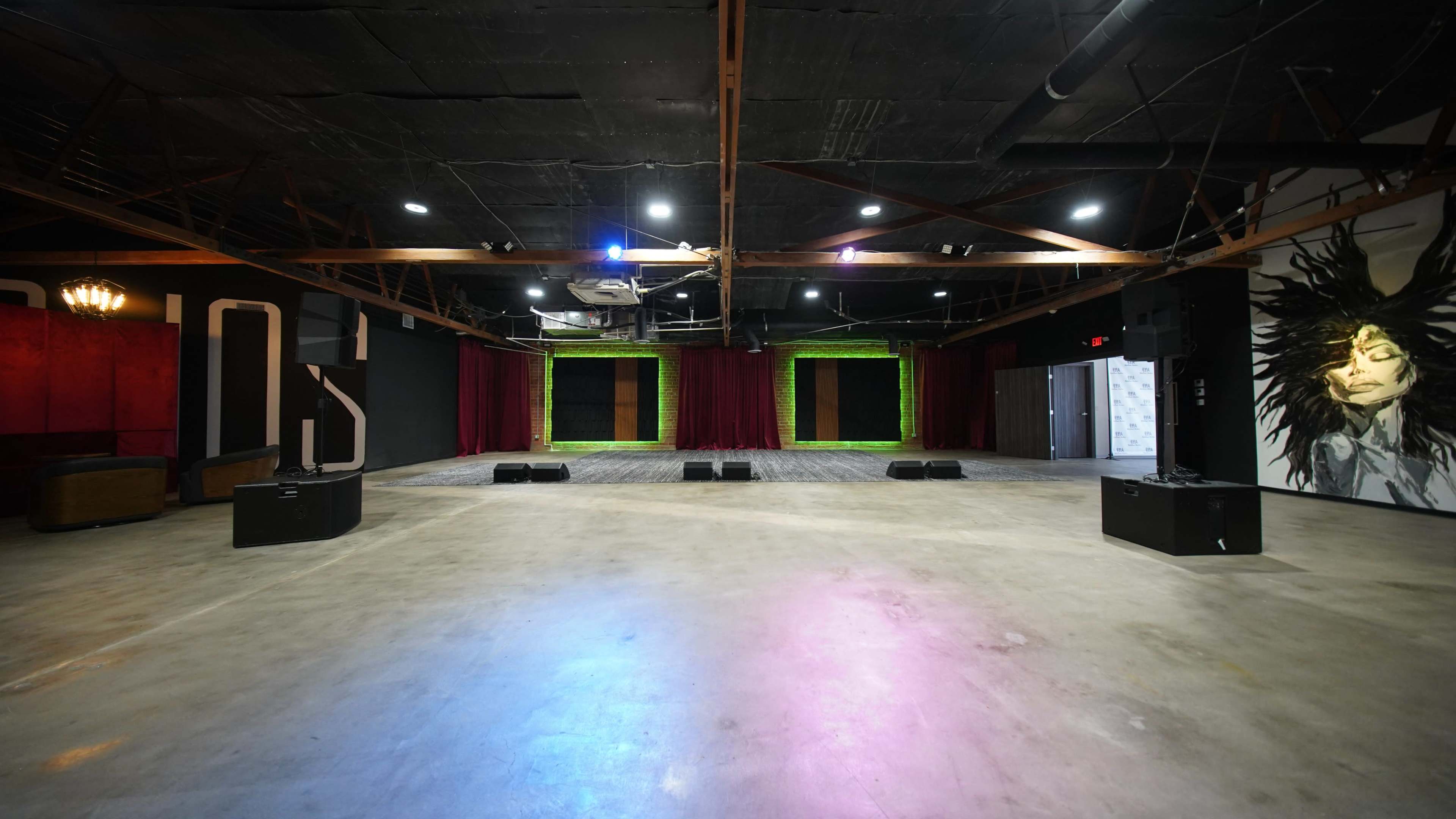 The image shows a spacious performance venue with a polished concrete floor, a stage at the front with red curtains, and a large mural on one wall.