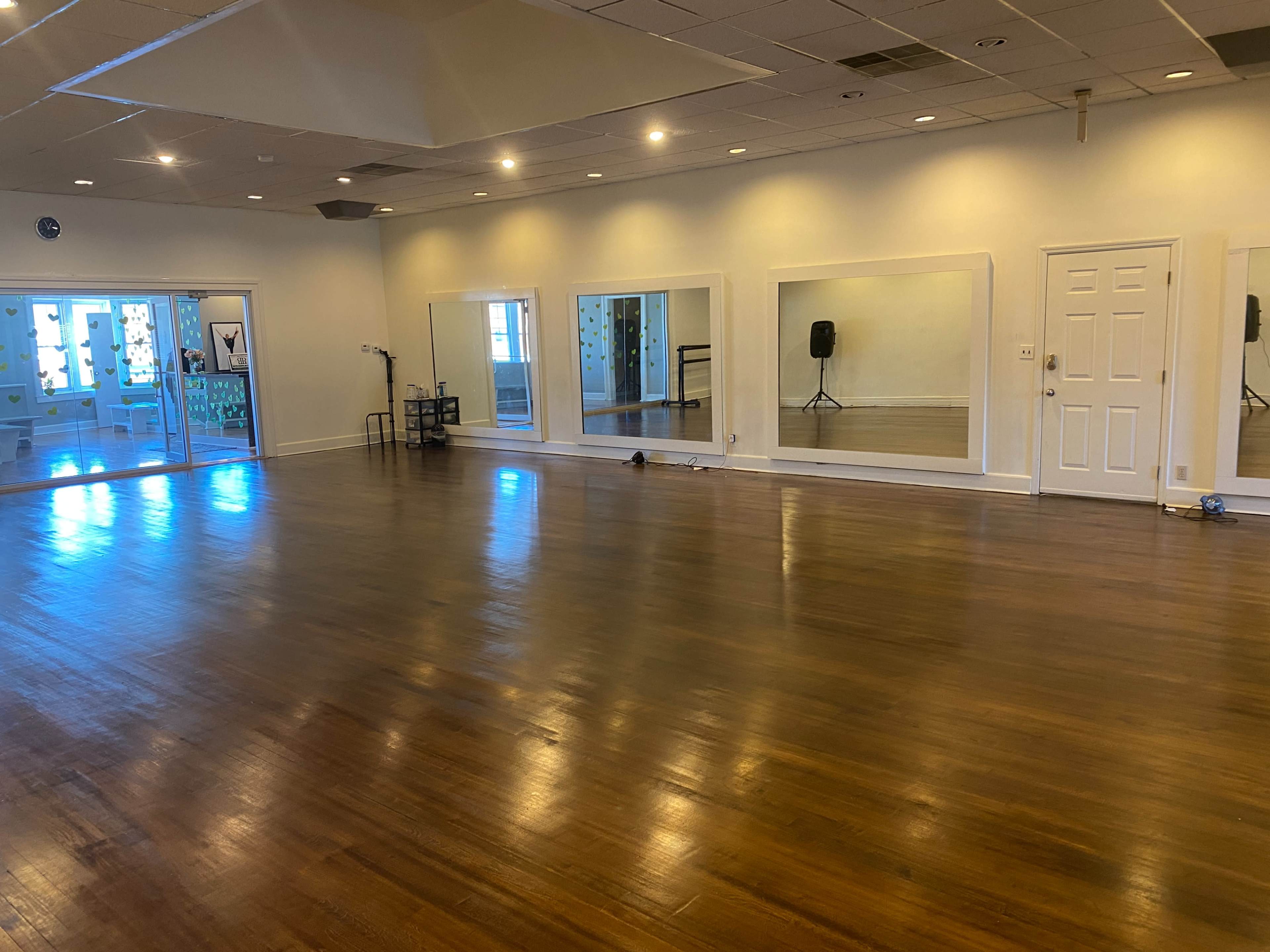 The image shows a spacious dance studio with polished wooden floors, large mirrors along one wall, and a door leading to an outdoor area.