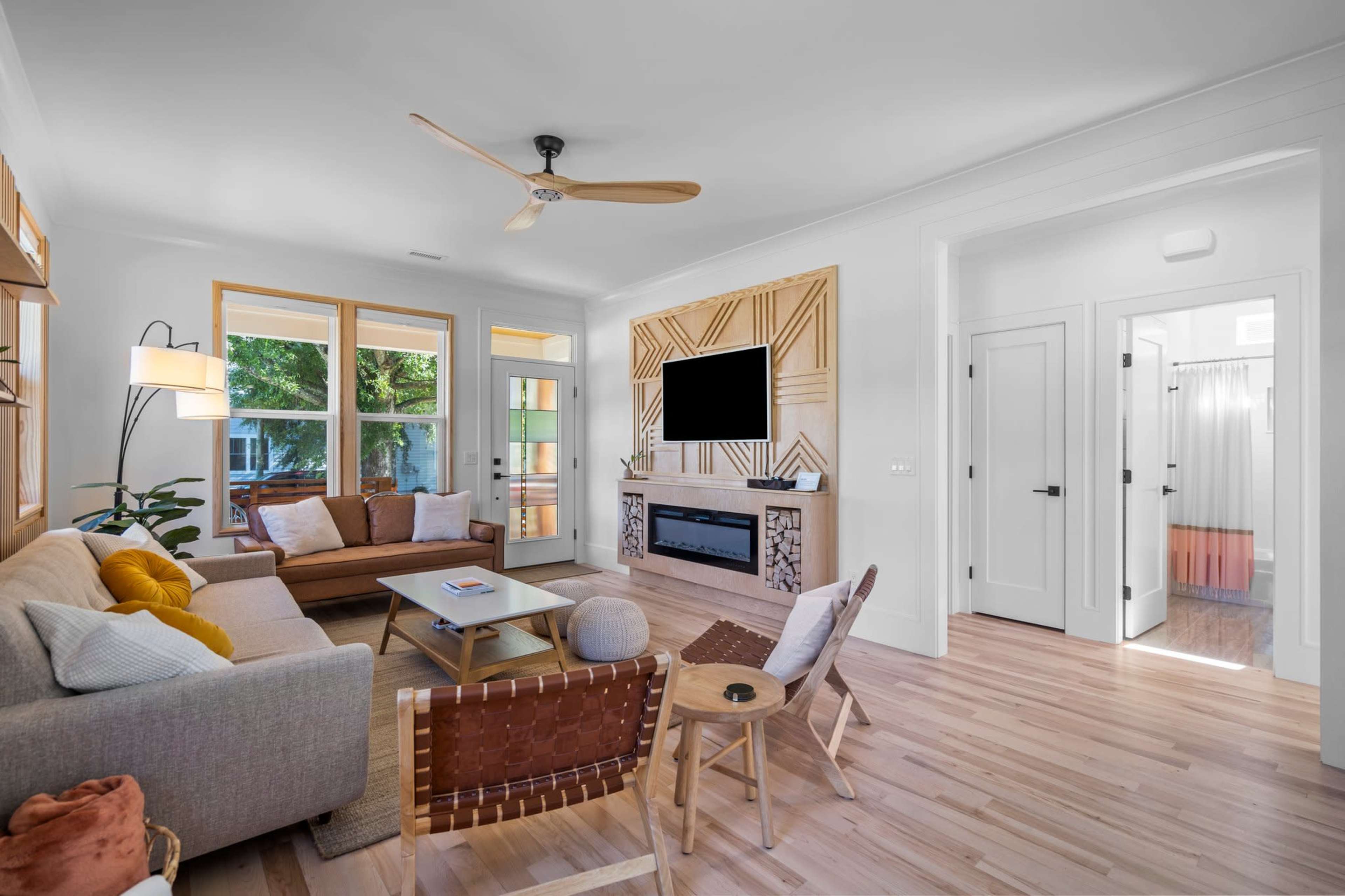 The living room features a sectional sofa, a coffee table, a wall-mounted TV, and large windows allowing natural light, with an adjacent hallway leading to another room.