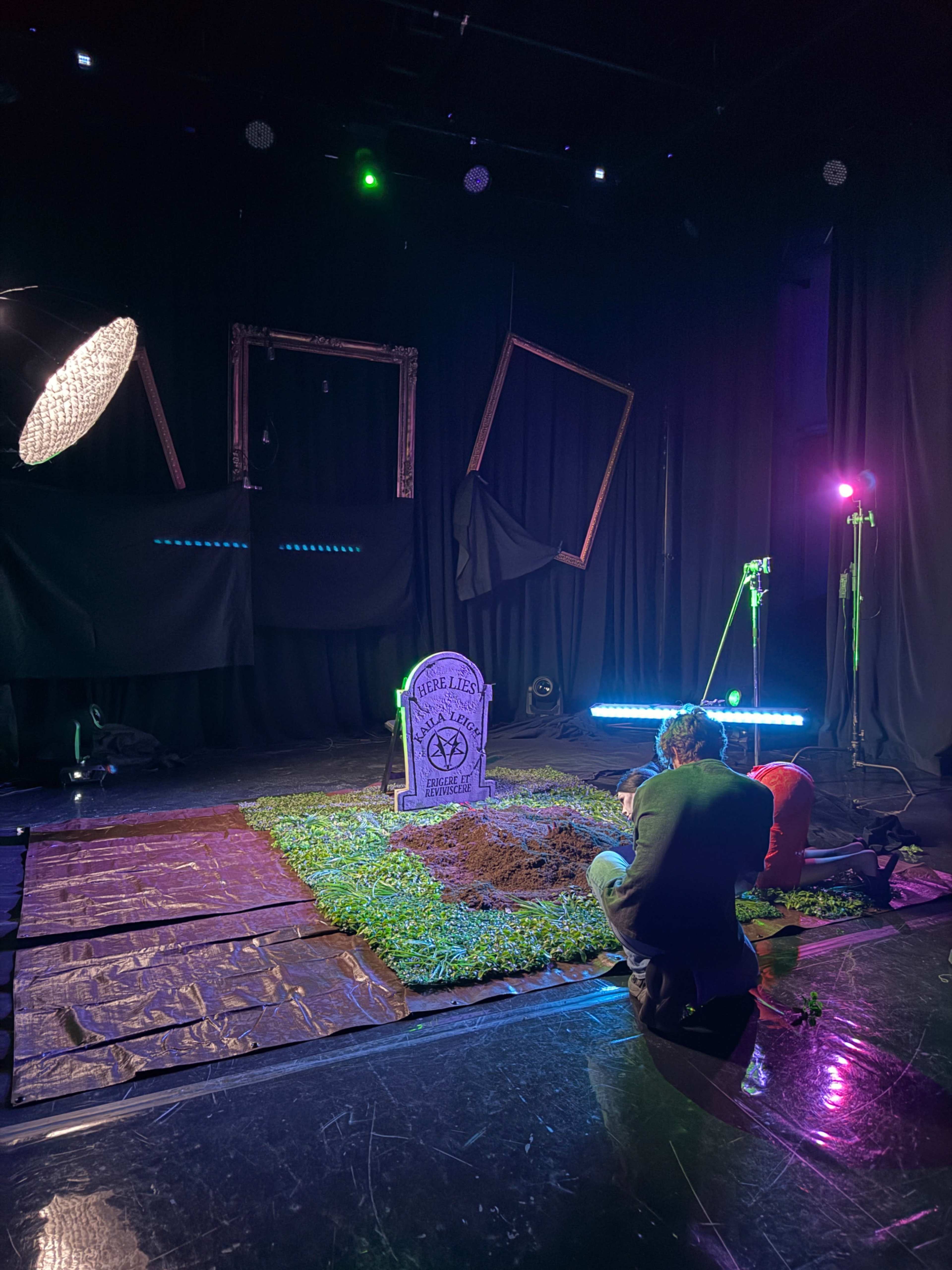A man sits on the floor in a dimly lit studio, focused on a tombstone labeled "RIP" surrounded by artificial grass and illuminated by colorful lights.