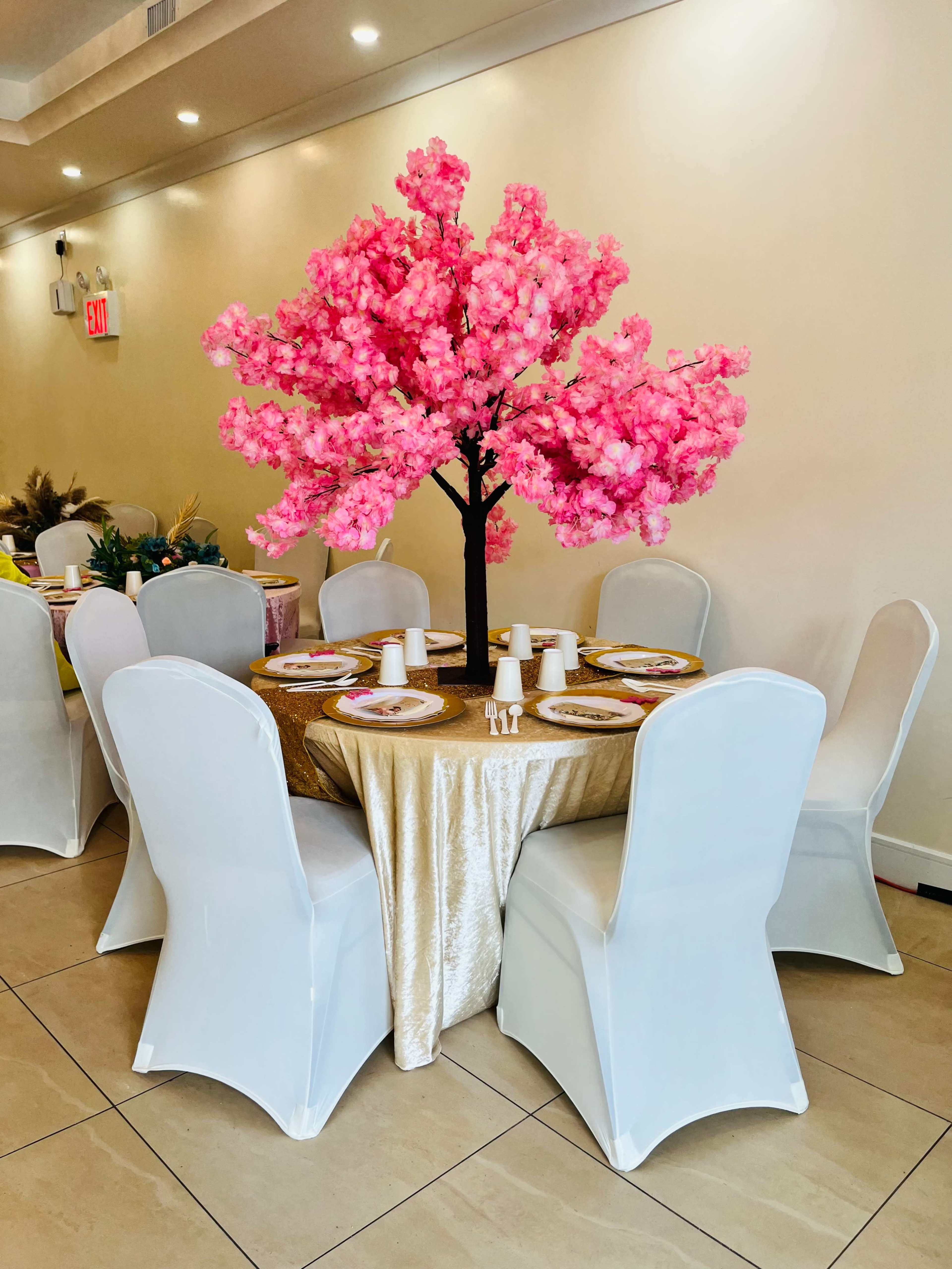 A round table is set with plates and cups, adorned with a large, pink flowering tree centerpiece in a banquet room.