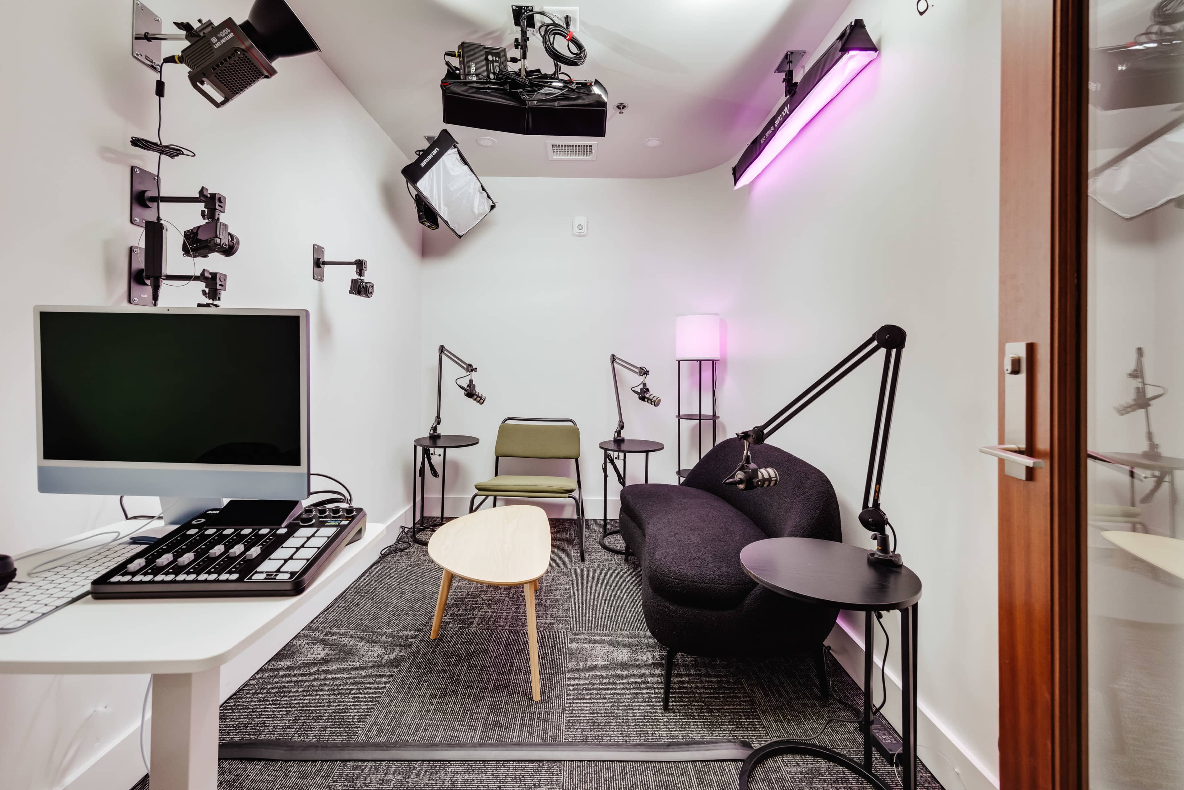 The image shows a small, modern recording studio with a computer, a cozy couch, a chair, and multiple mounted microphones.