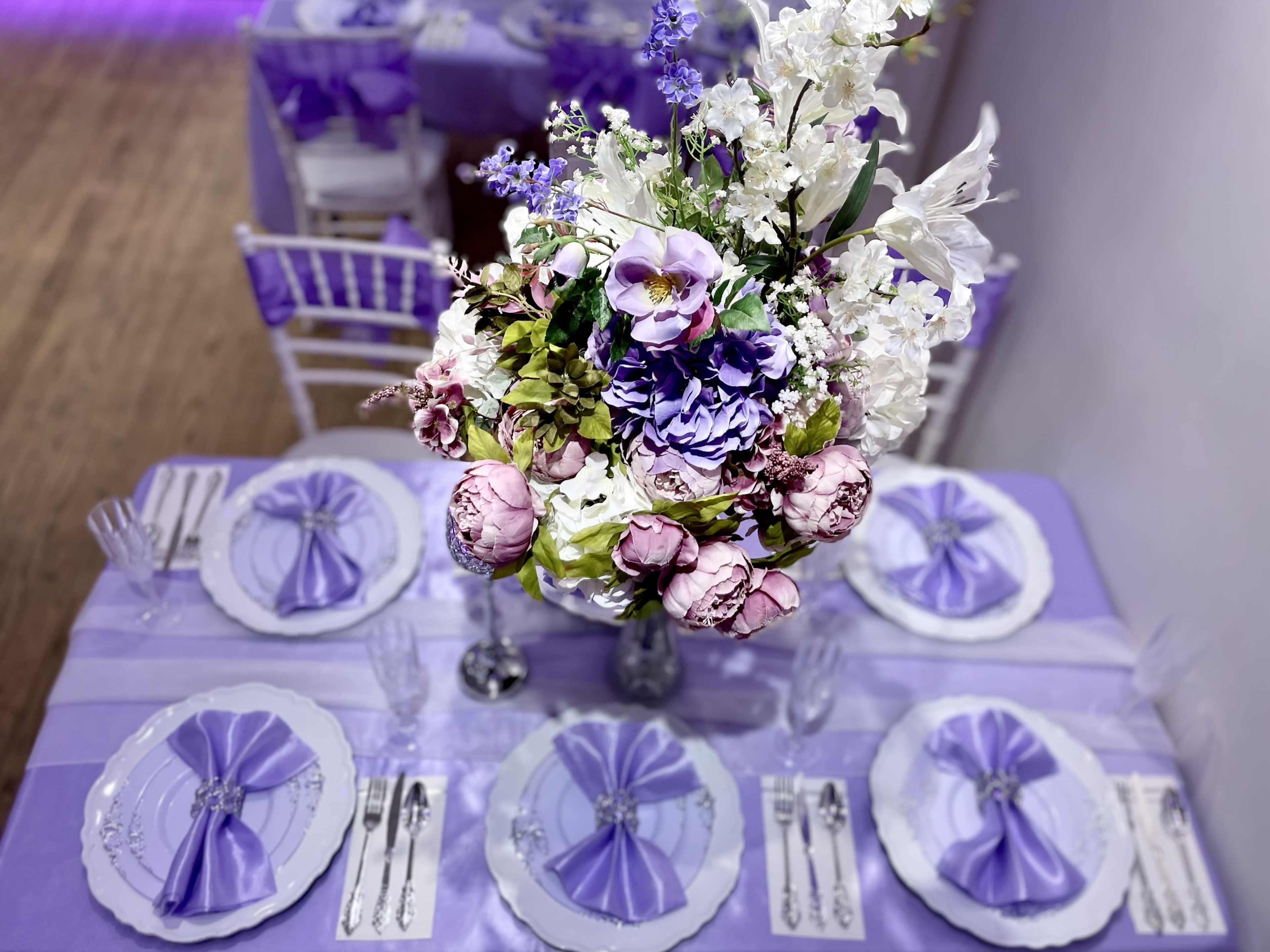 A table set with purple and white decorations features an ornate floral centerpiece and intricately folded napkins.