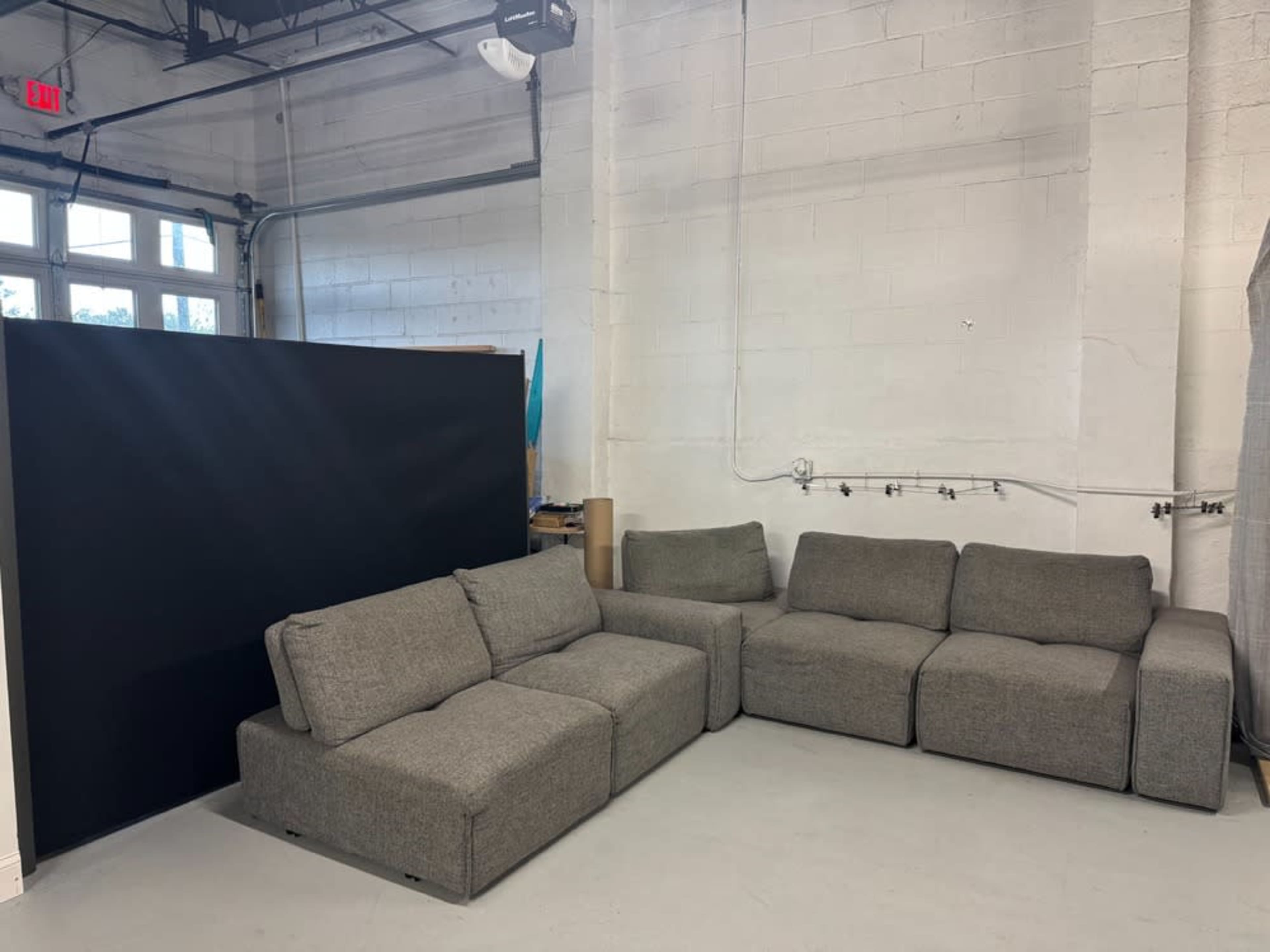 The image shows a gray sectional sofa against a white wall in a sparsely furnished indoor space.