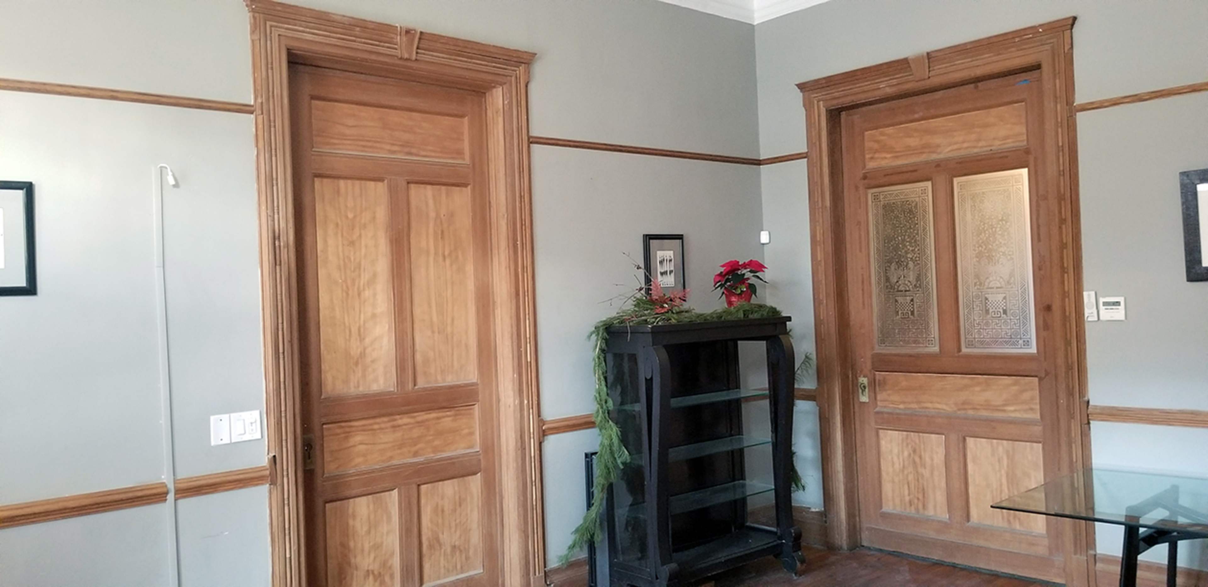 The image shows a room with two wooden doors, one plain and one with decorative glass panels, alongside a black bookcase and a glass table against grey walls.