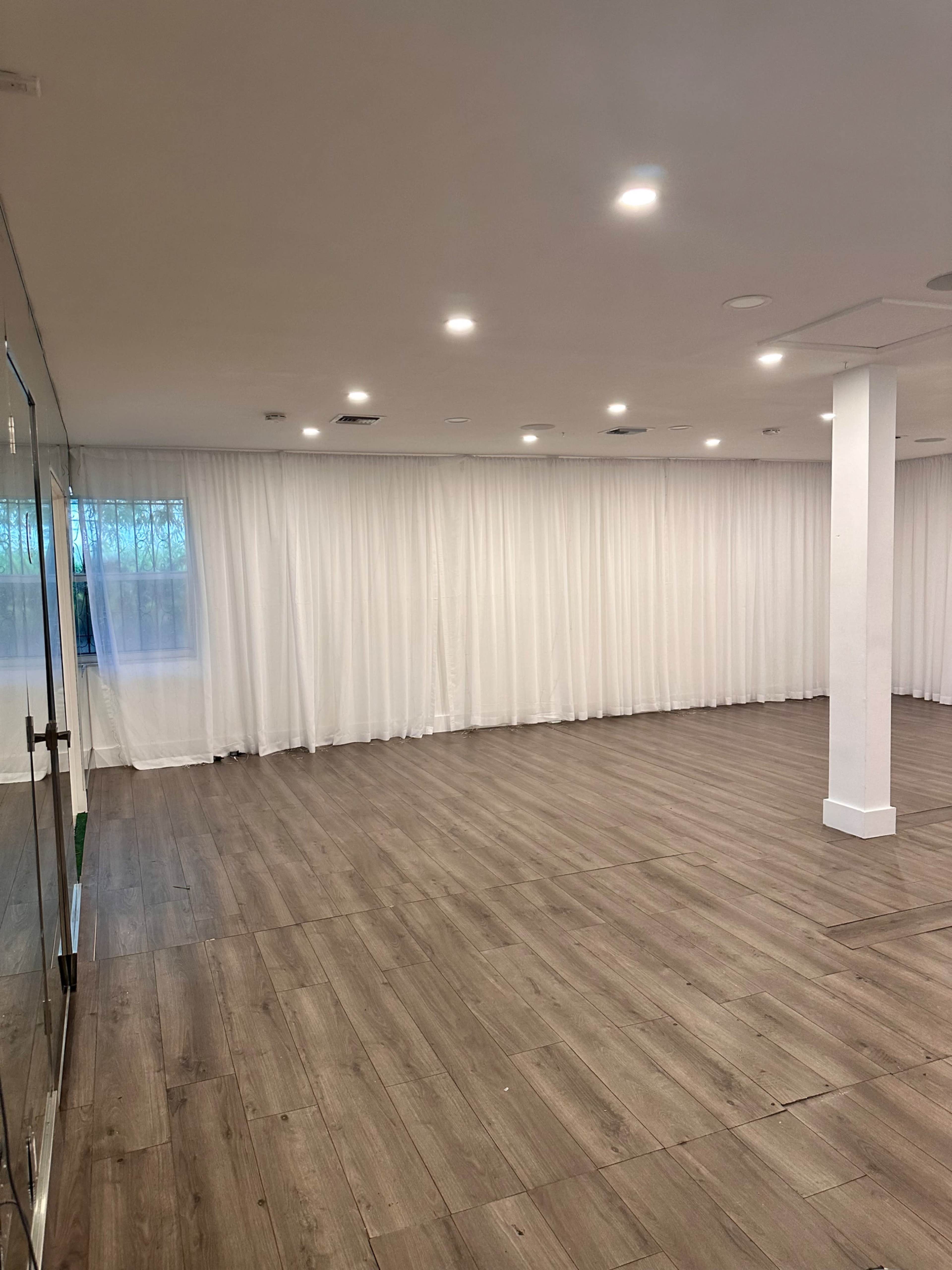 An empty room with a polished wooden floor, white curtains along one wall, and recessed lighting.