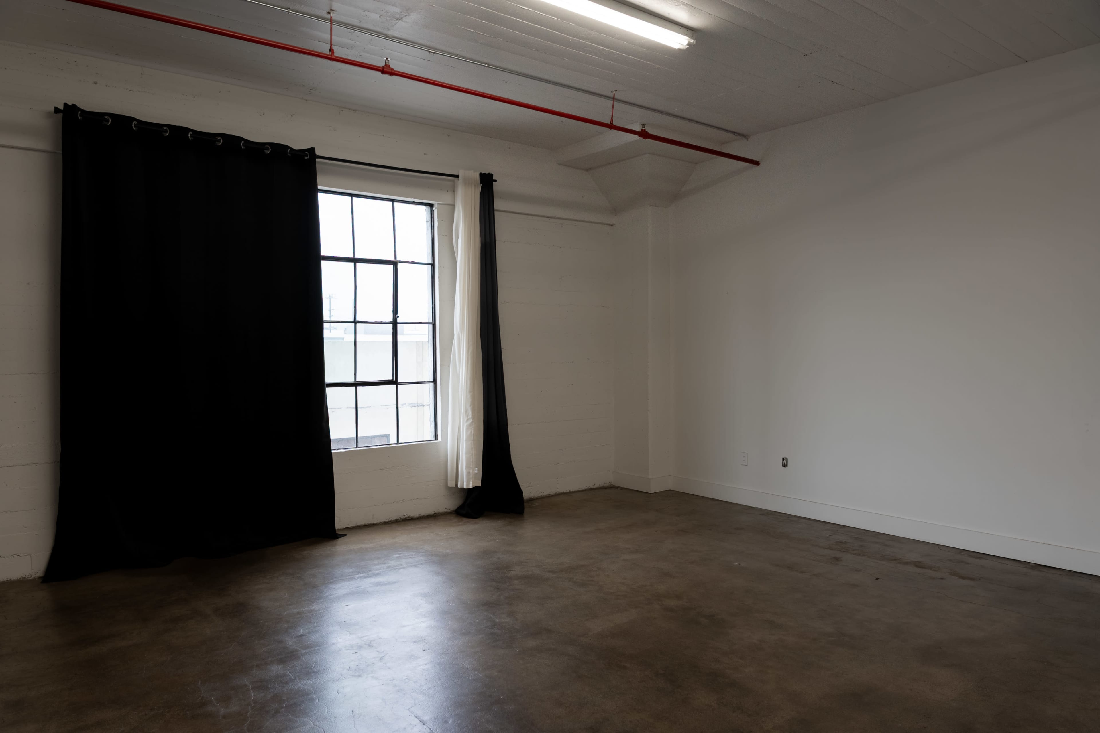 The image shows a sparsely furnished room with a large window covered by black and white curtains and concrete flooring.
