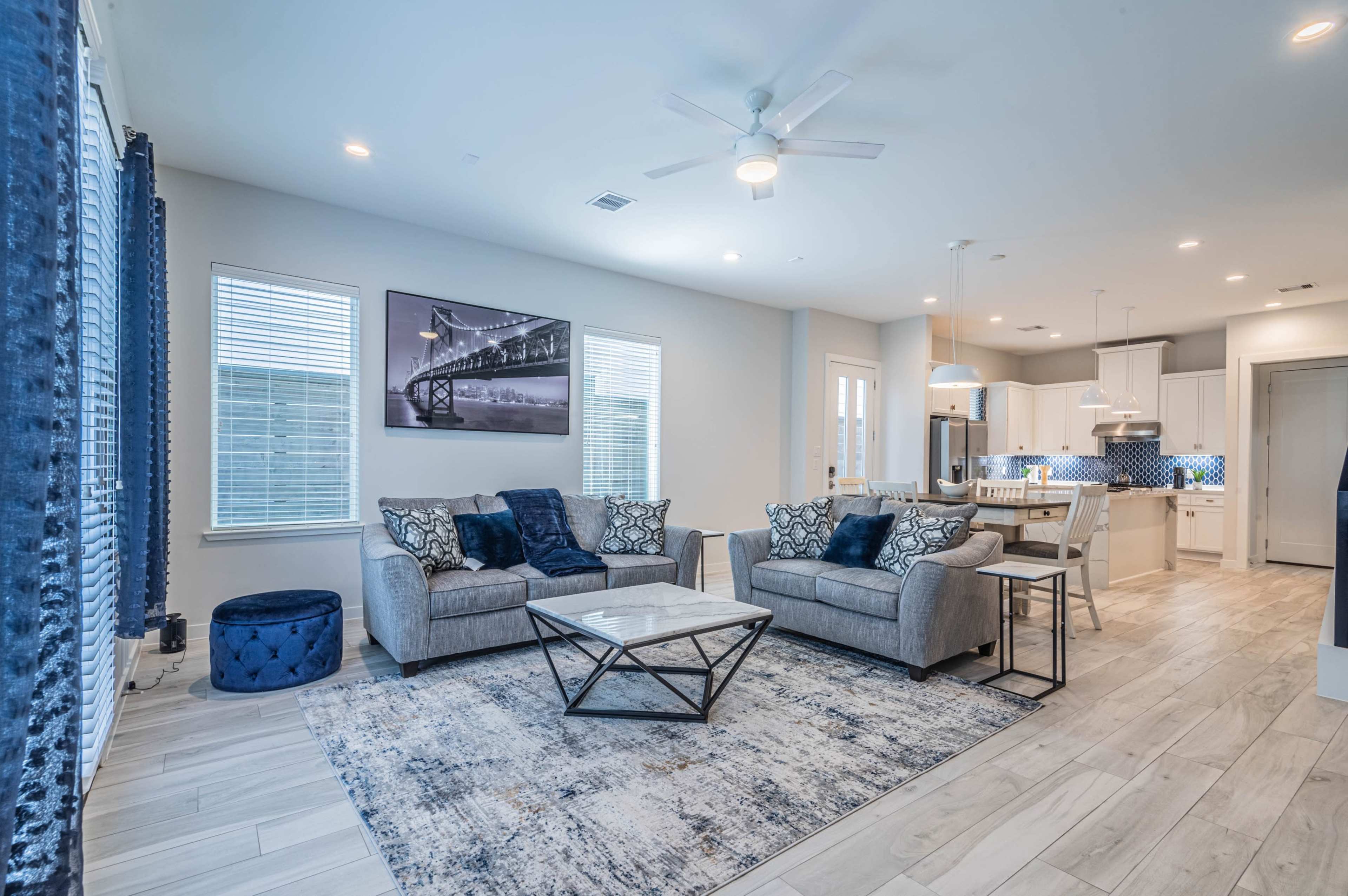 A modern living room with gray sofas, a coffee table, and a dining area visible in the background, all framed by blue decor and well-lit windows.