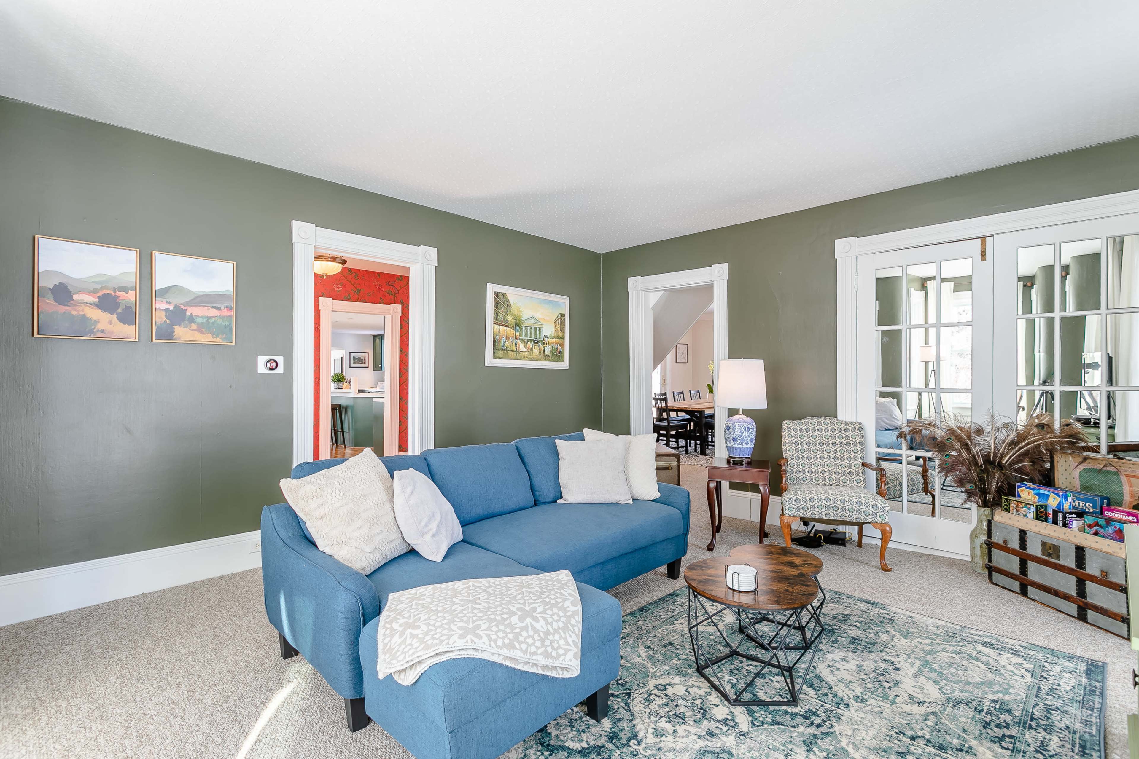 A blue sofa with decorative pillows sits in a living room featuring green walls, a patterned area rug, and a coffee table, with doors leading to another room.