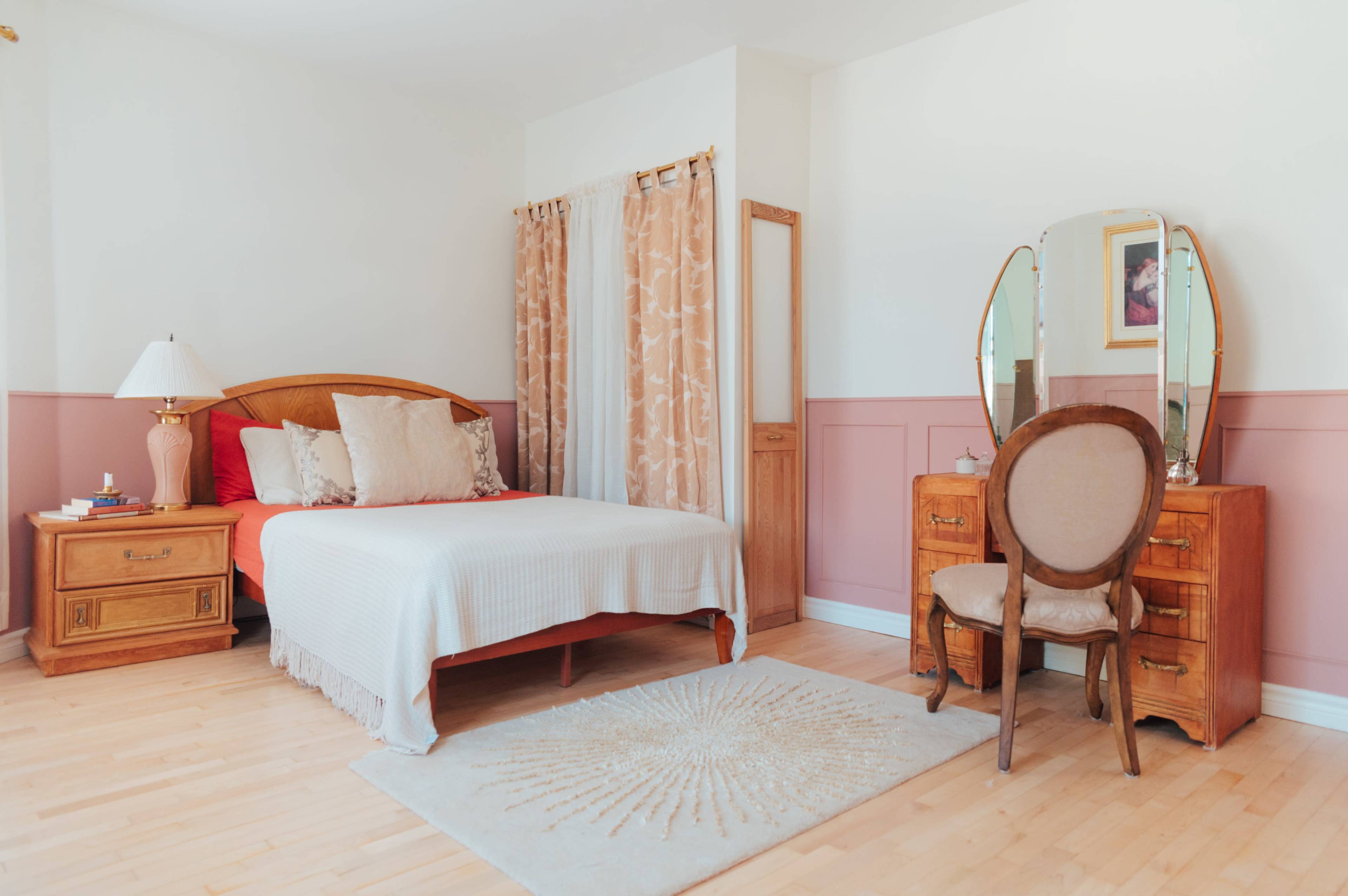 The image shows a bedroom featuring a bed with a white bedspread, two wooden nightstands, a mirrored dresser, and a chair, all set against a light-colored wall with decorative curtains.