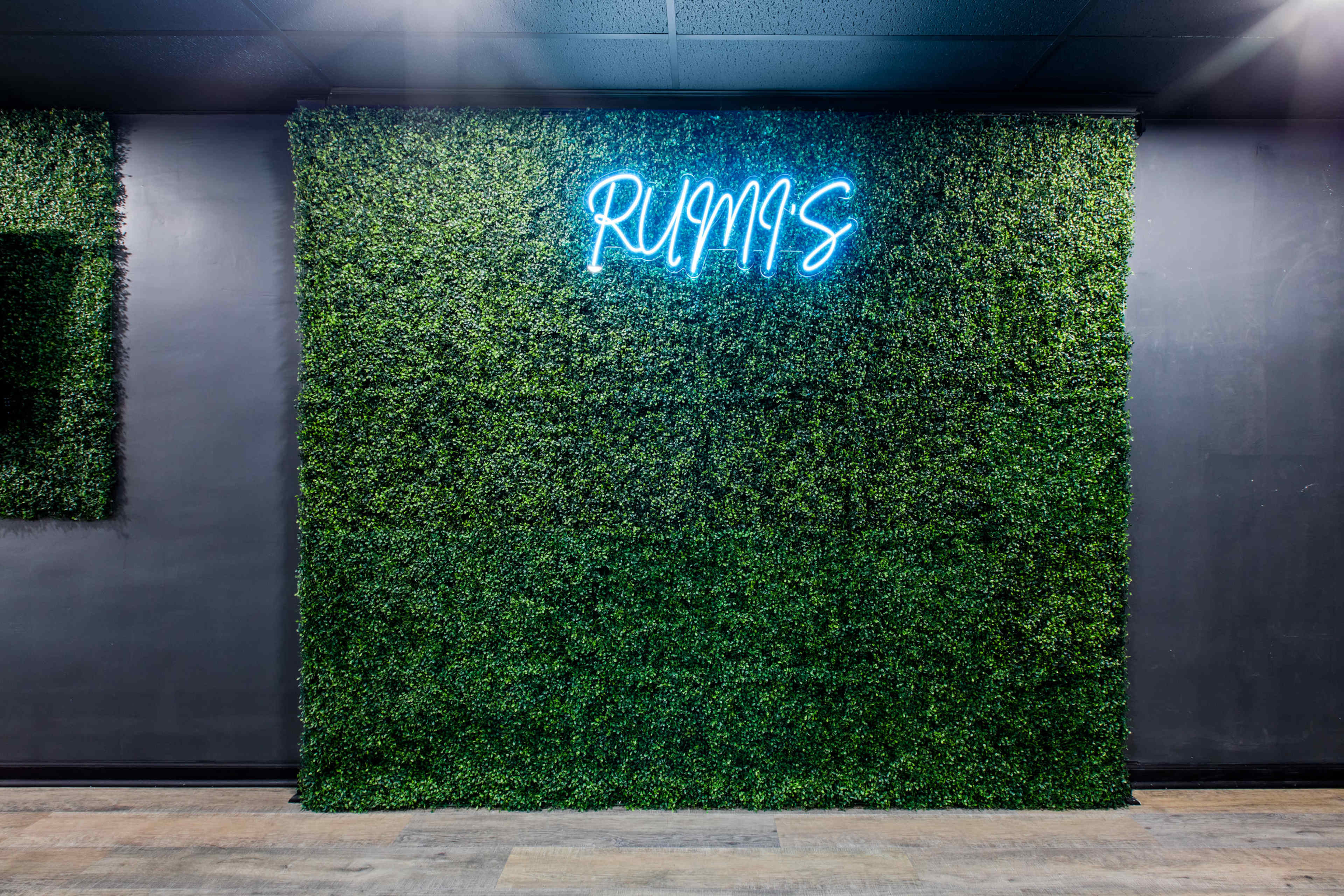 A wall covered in artificial greenery features a neon sign that reads "RUMIS."