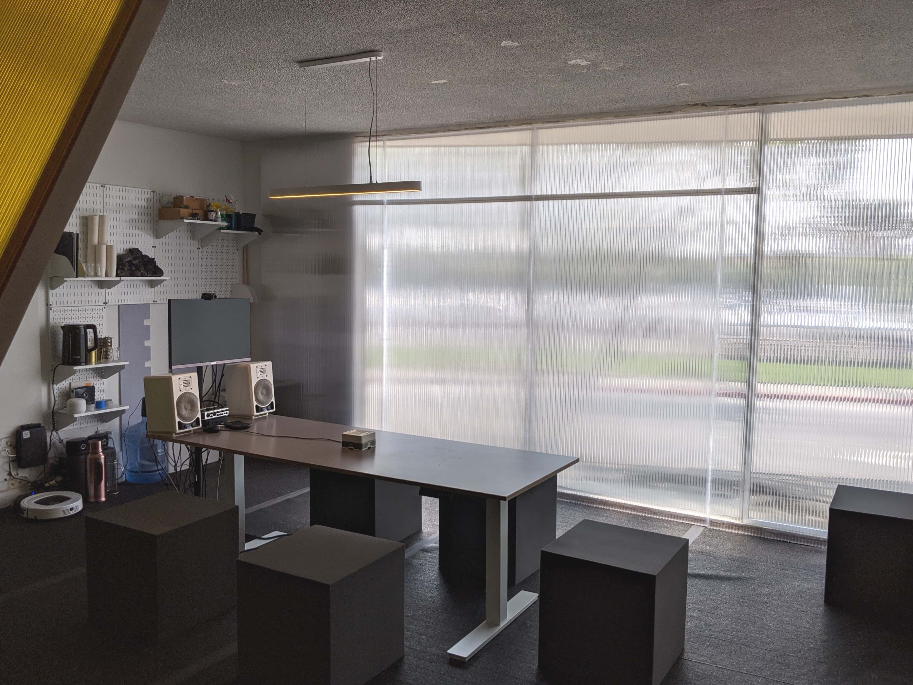 A minimalist meeting room features a table, two speakers, and several square stools, with large windows allowing natural light to enter.