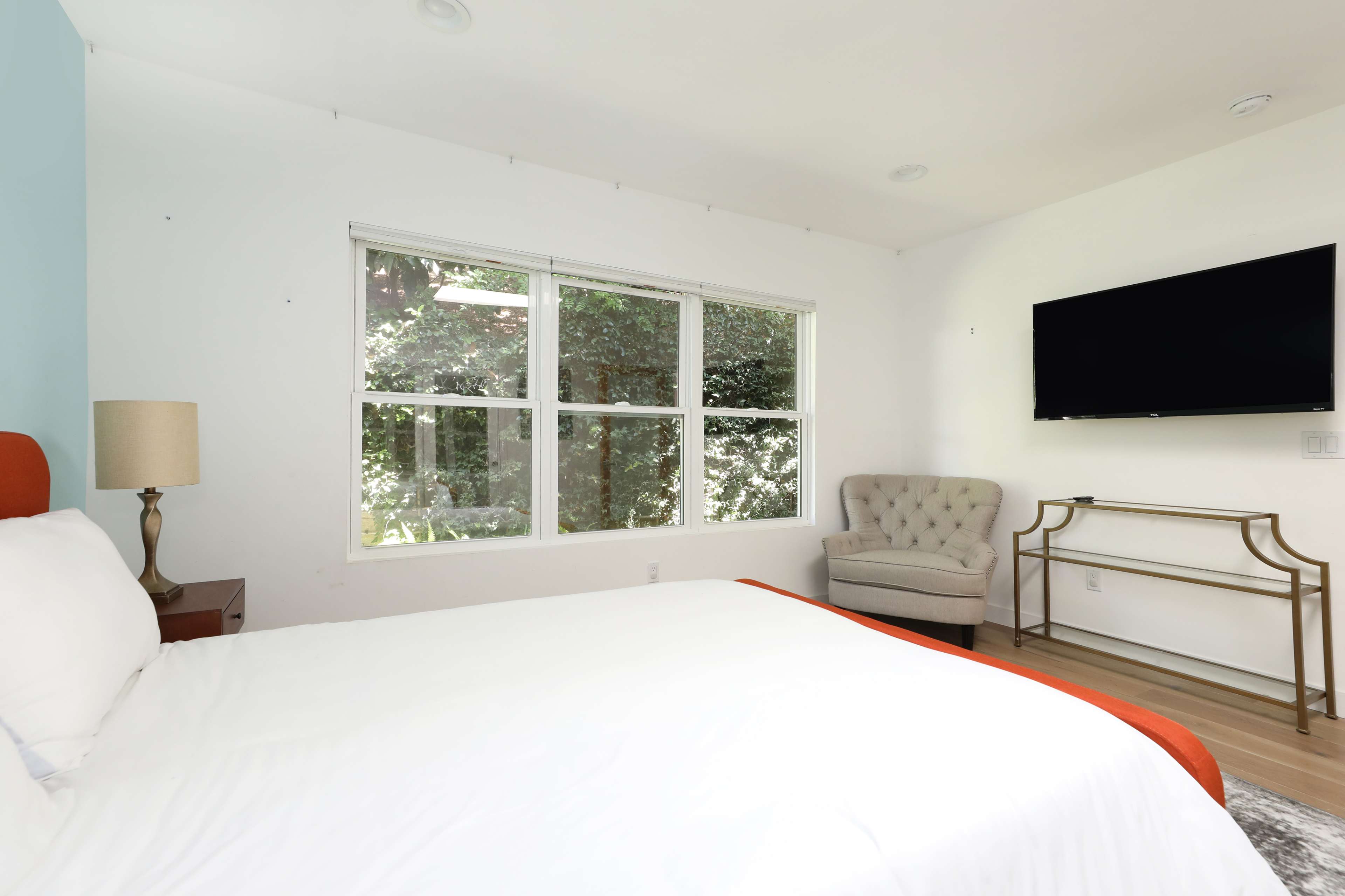 The image shows a bedroom featuring a bed with a white blanket, a tufted armchair, a lamp, a wall-mounted television, and large windows with views of greenery outside.
