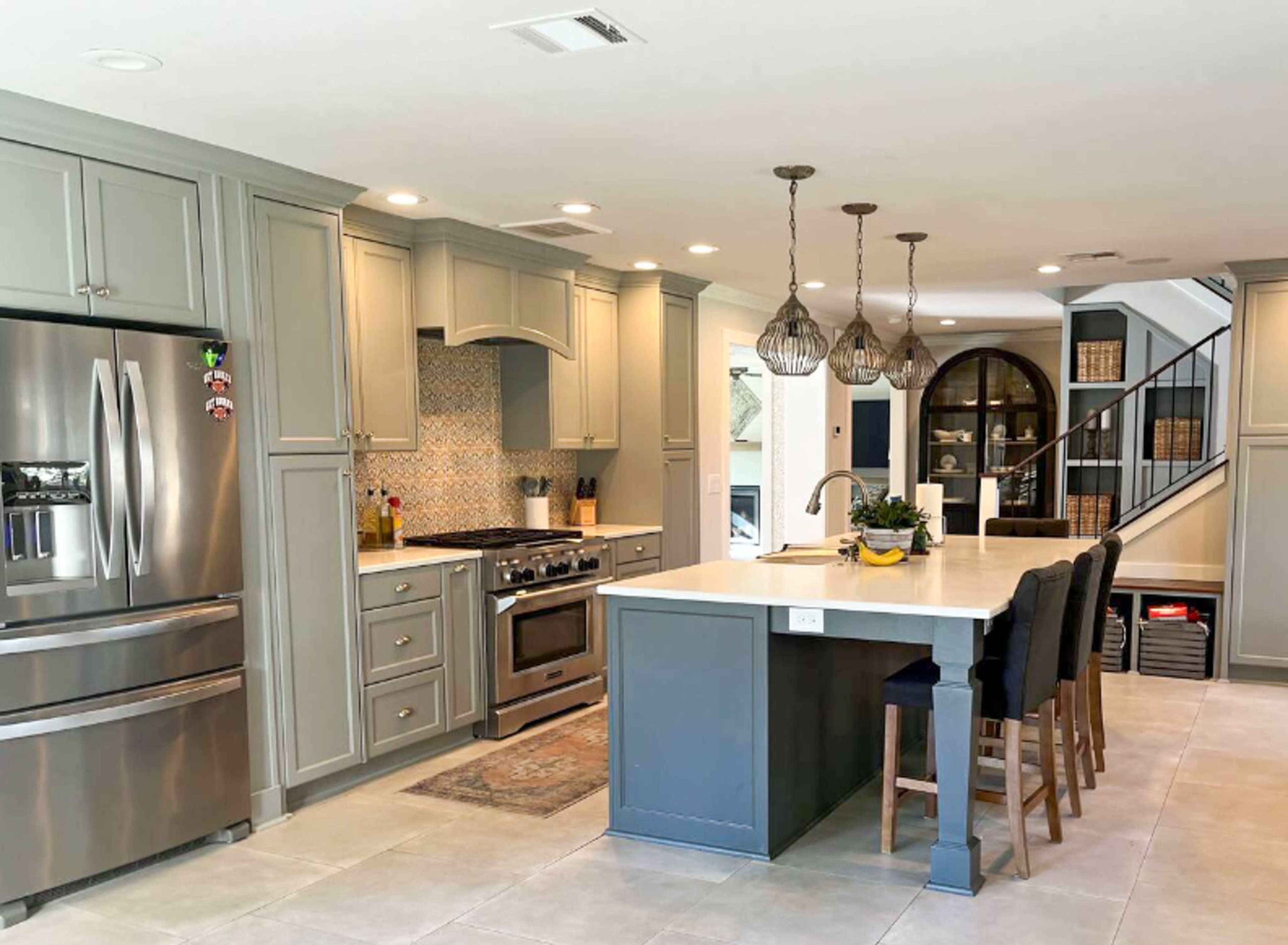The image shows a modern kitchen with gray cabinetry, stainless steel appliances, and an island with seating, leading to a staircase in the background.