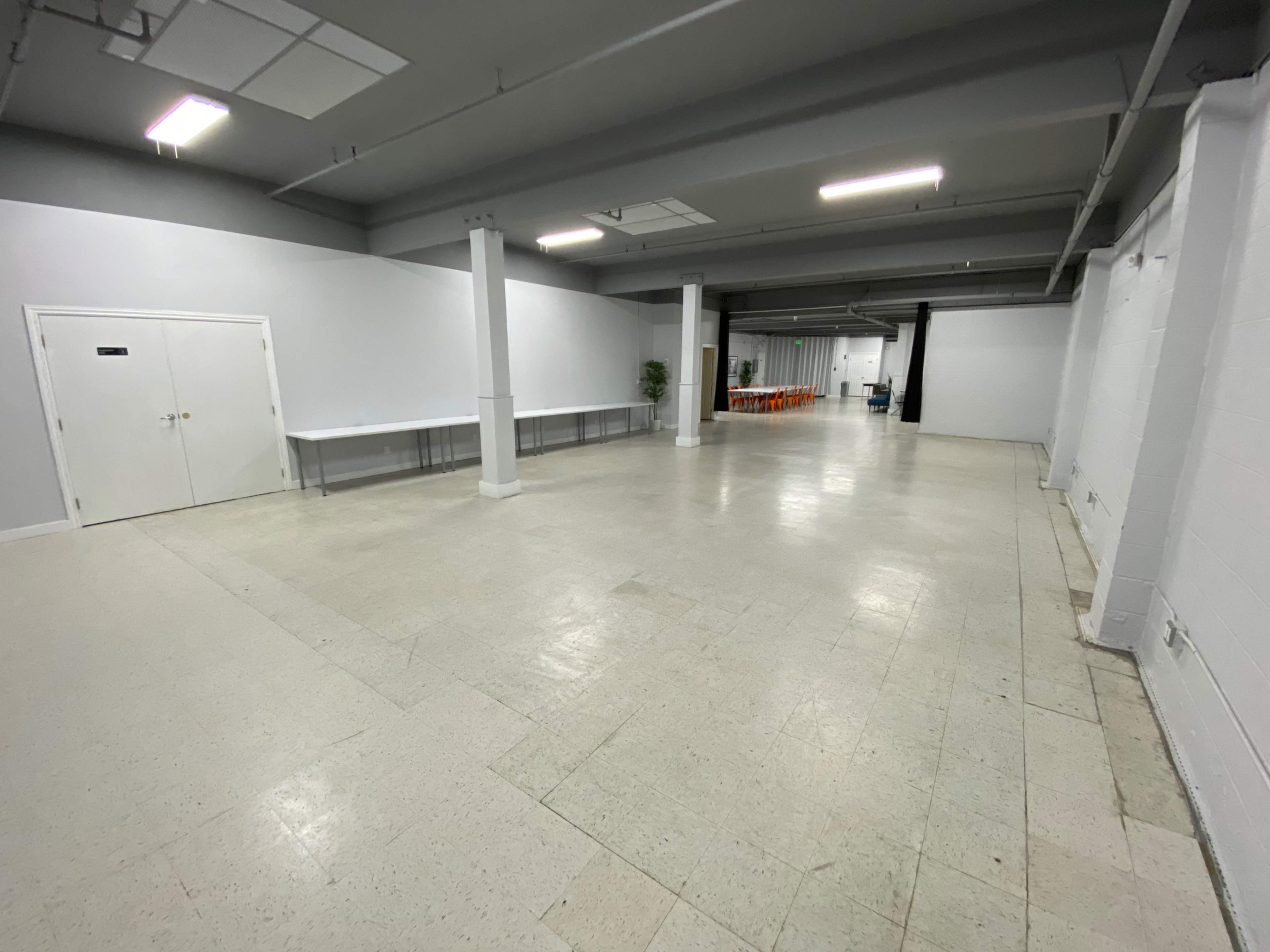 A spacious, mostly empty room with white walls, a concrete floor, and several tables set against one side.