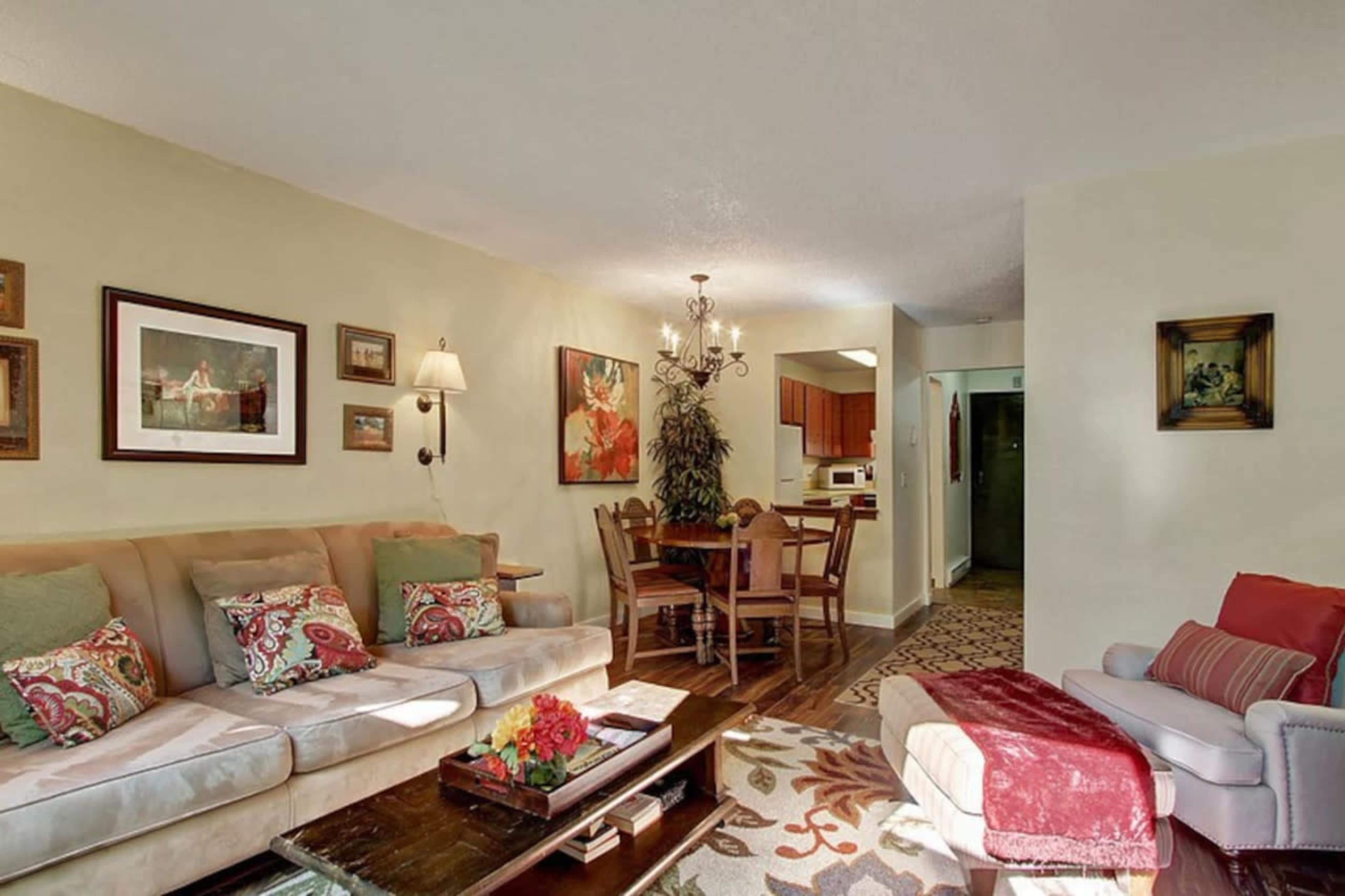 The image shows a living room with a beige couch, decorative pillows, a dining area with wooden chairs, and wall art, all in a well-lit space.