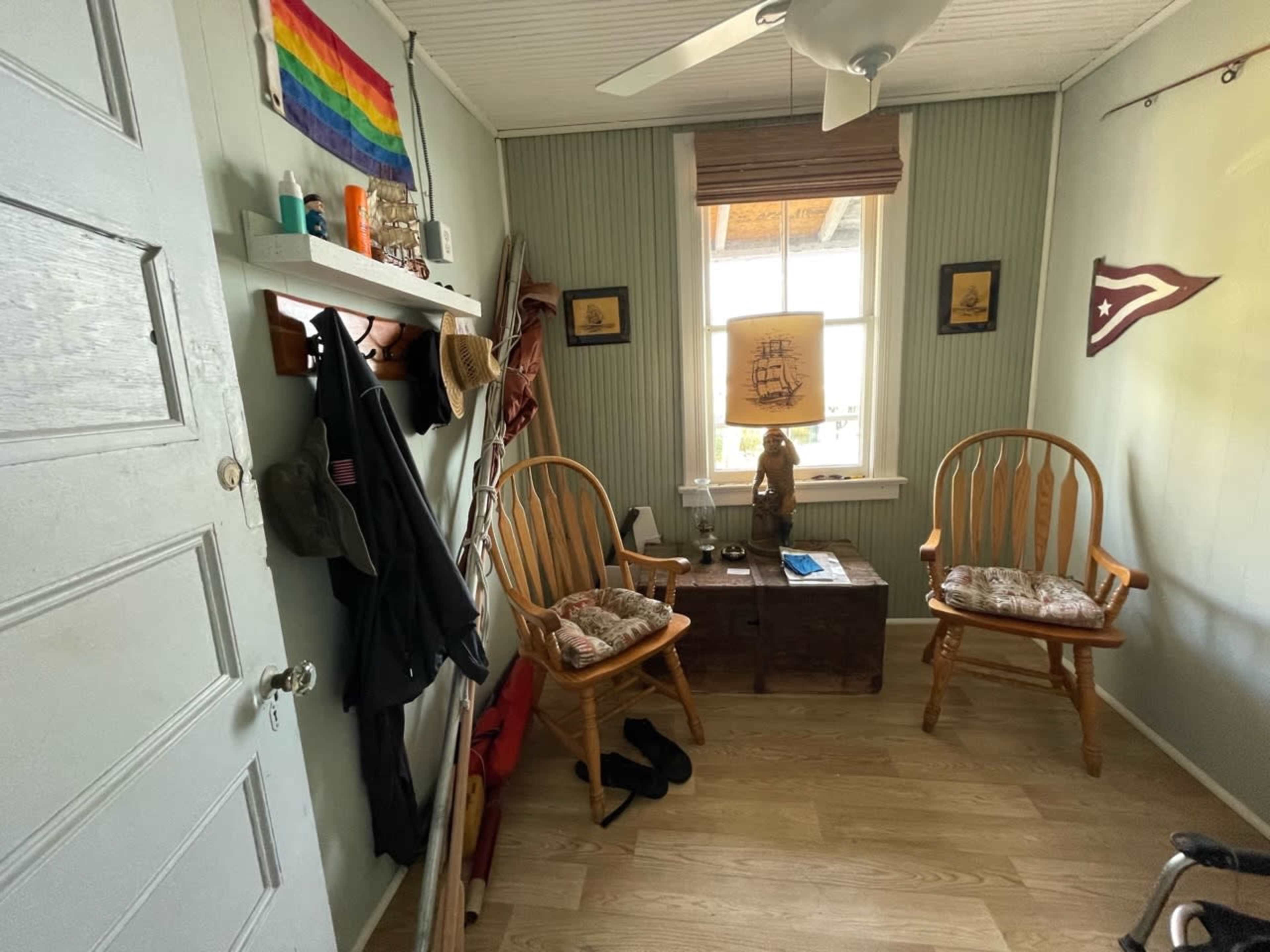 A small room with two wooden chairs, a shelf with clothing items, and a window with a blind, featuring walls covered in green paneling and decorations such as flags and framed pictures.
