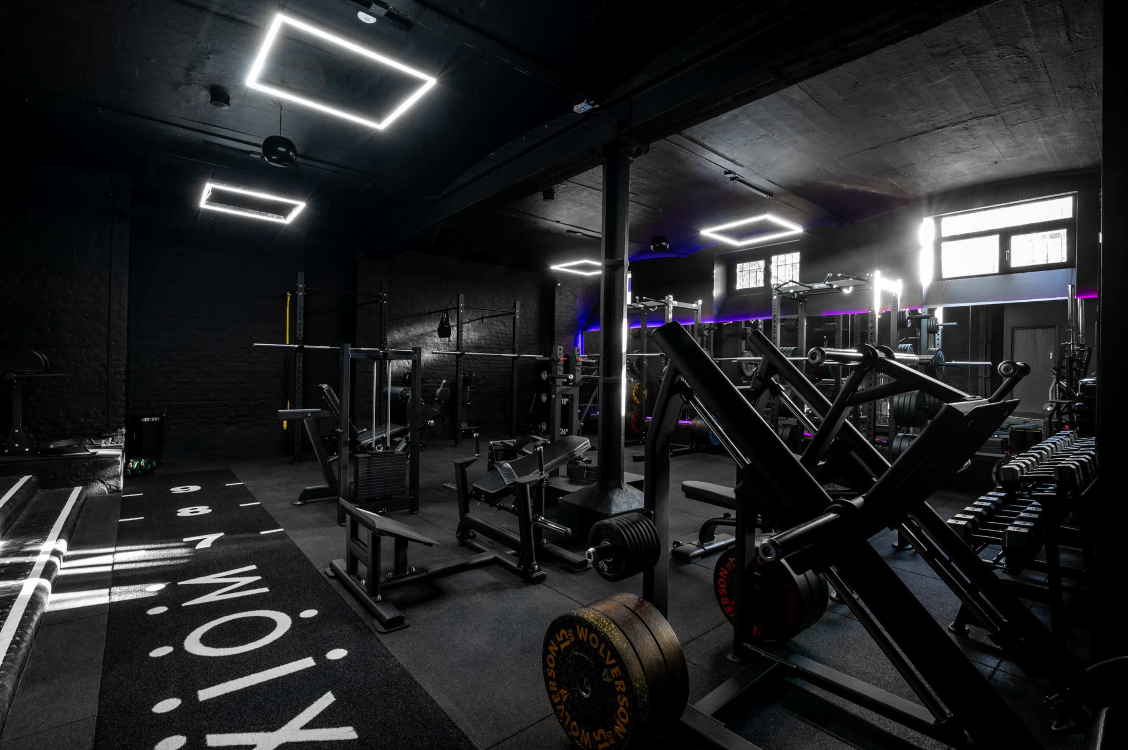 The image shows a dimly lit gym filled with various weight training equipment and illuminated by modern ceiling lights.