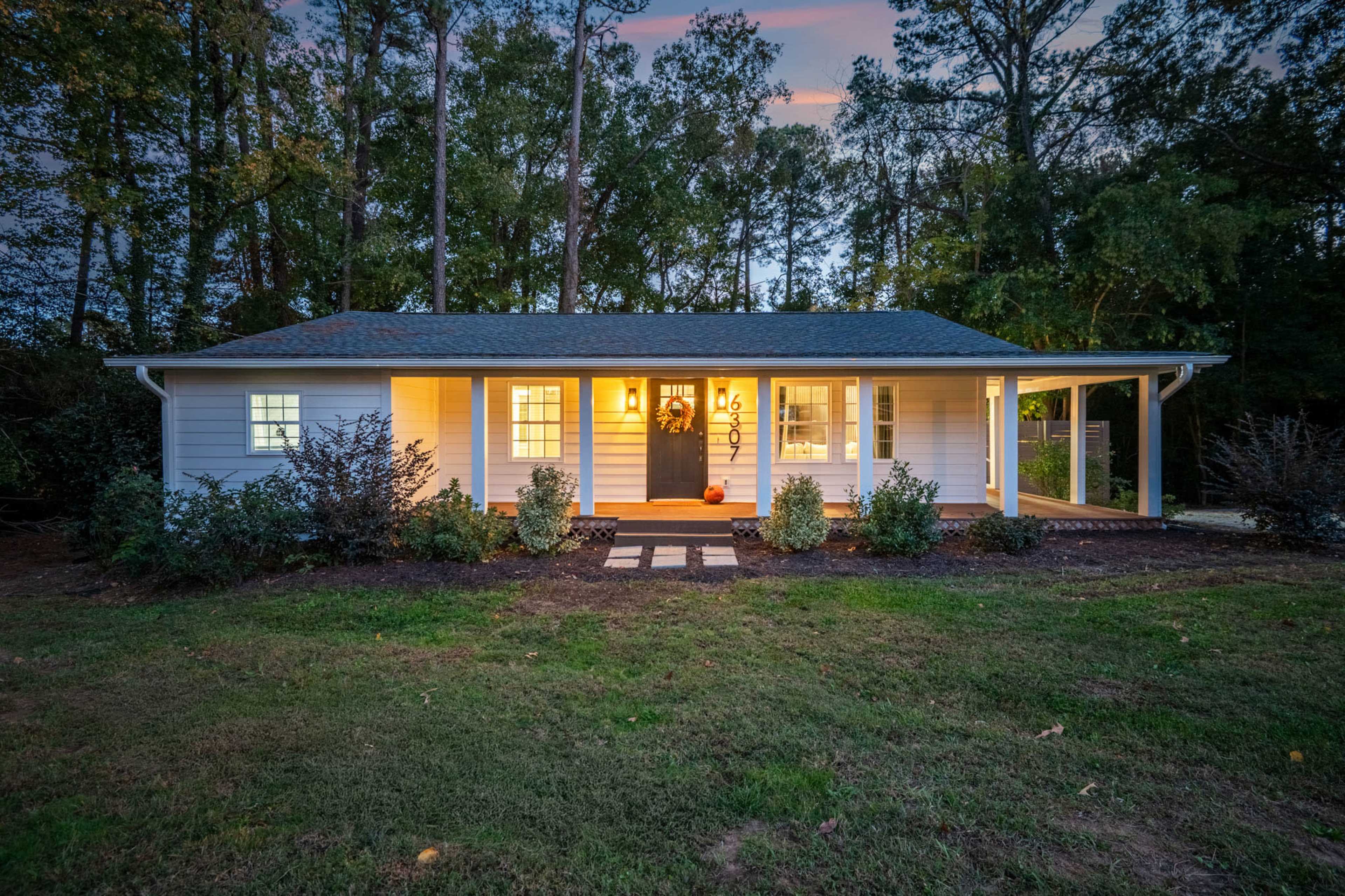 A single-story house with a porch is illuminated in the evening, surrounded by trees and landscaped grass.
