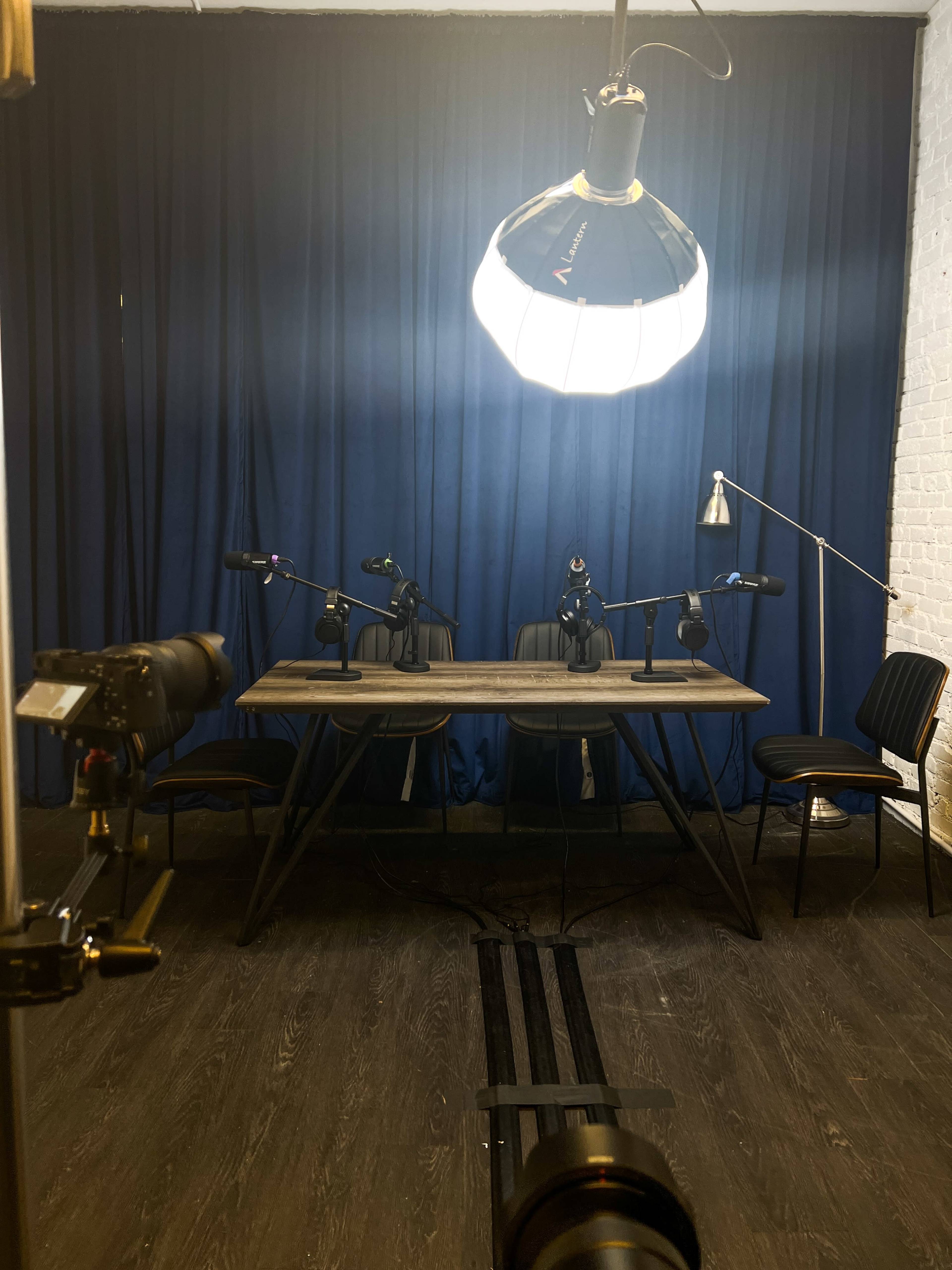 A podcast recording setup with a table, three microphones, a lamp, and a dark blue curtain in the background.