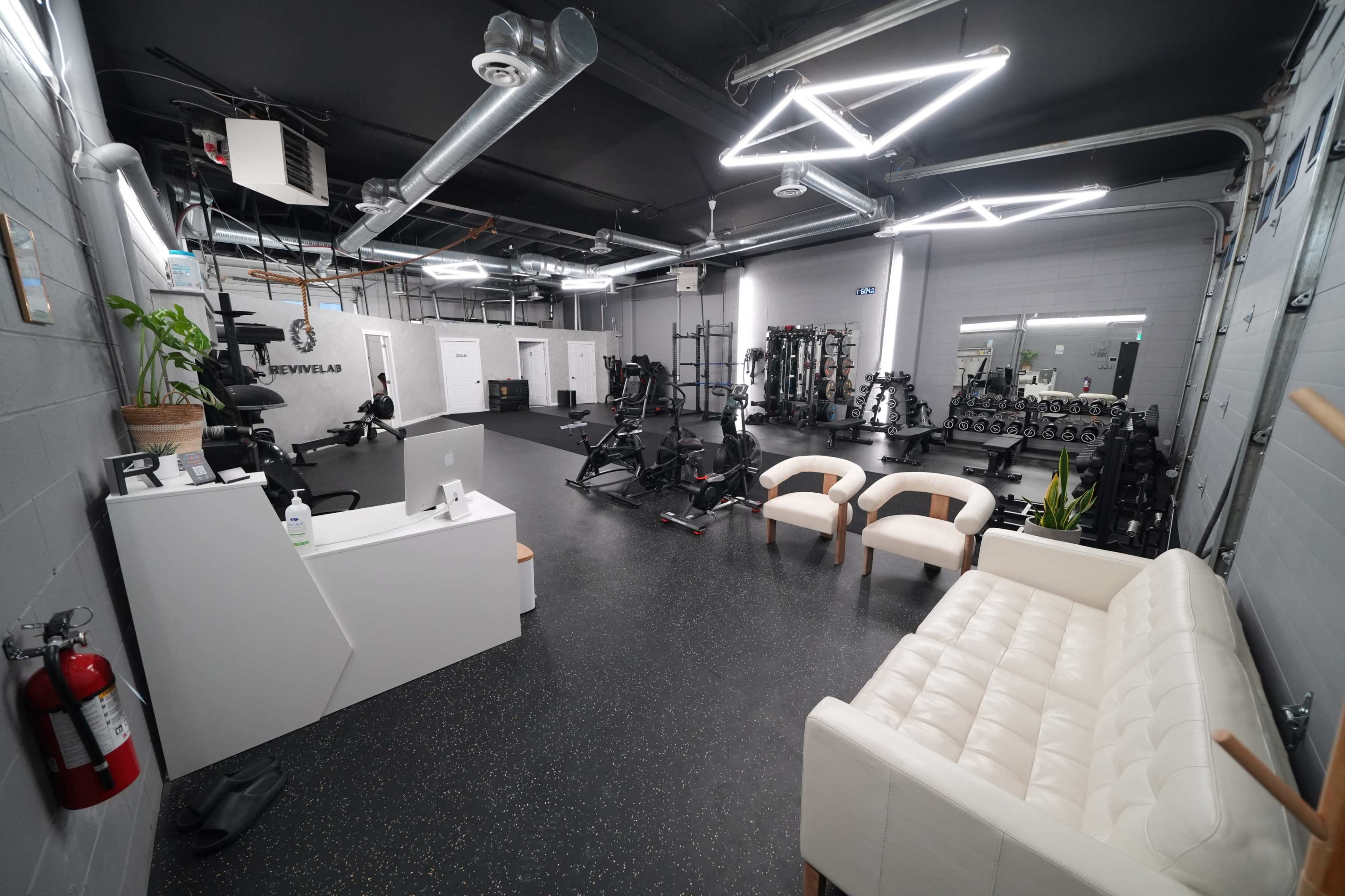The image shows a modern gym interior with various exercise equipment, a reception area featuring a white desk, and two beige chairs facing each other.