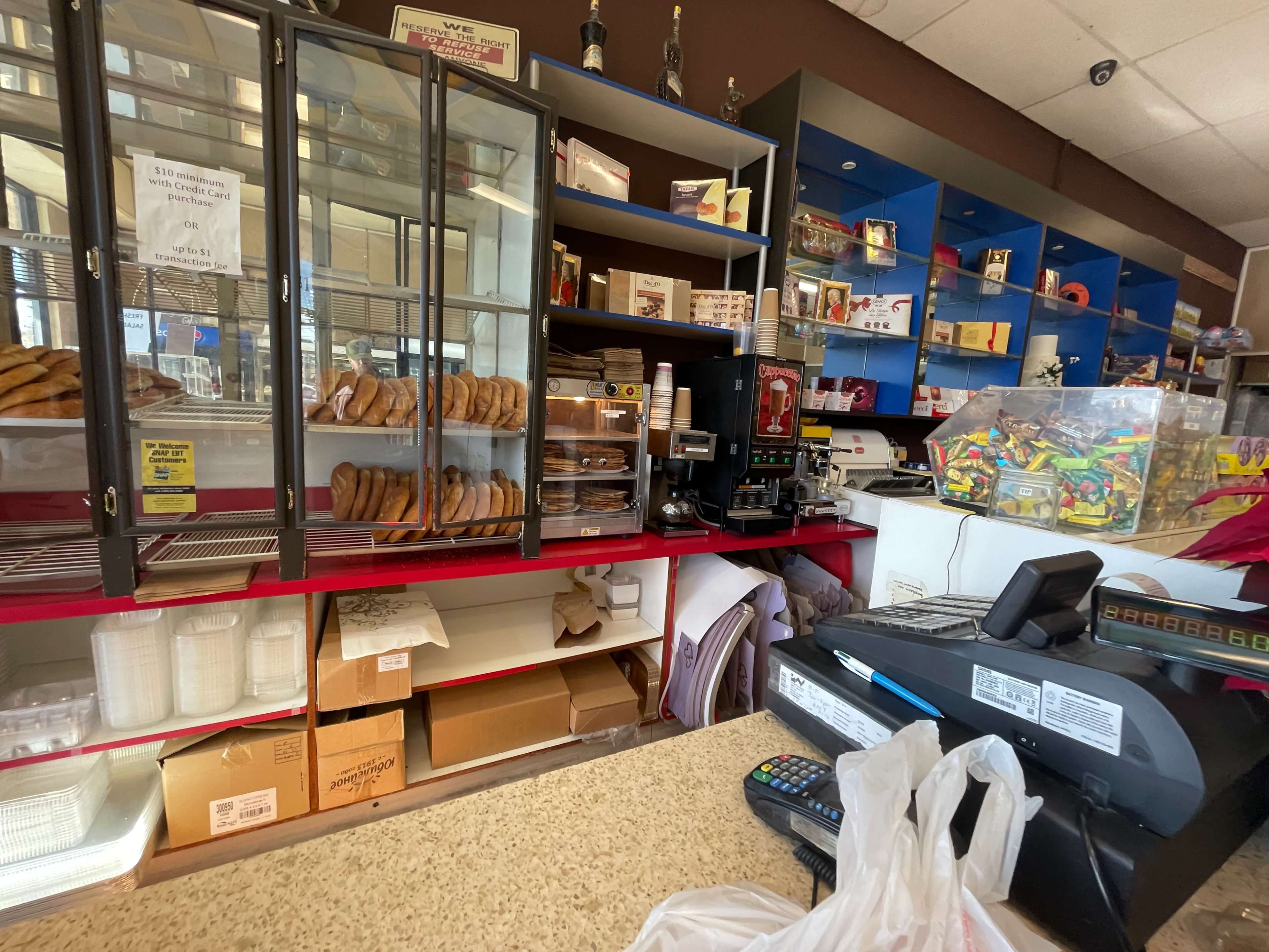 The image shows the interior of a bakery with a glass display case filled with baked goods, a cash register, and shelves lined with various snack items.