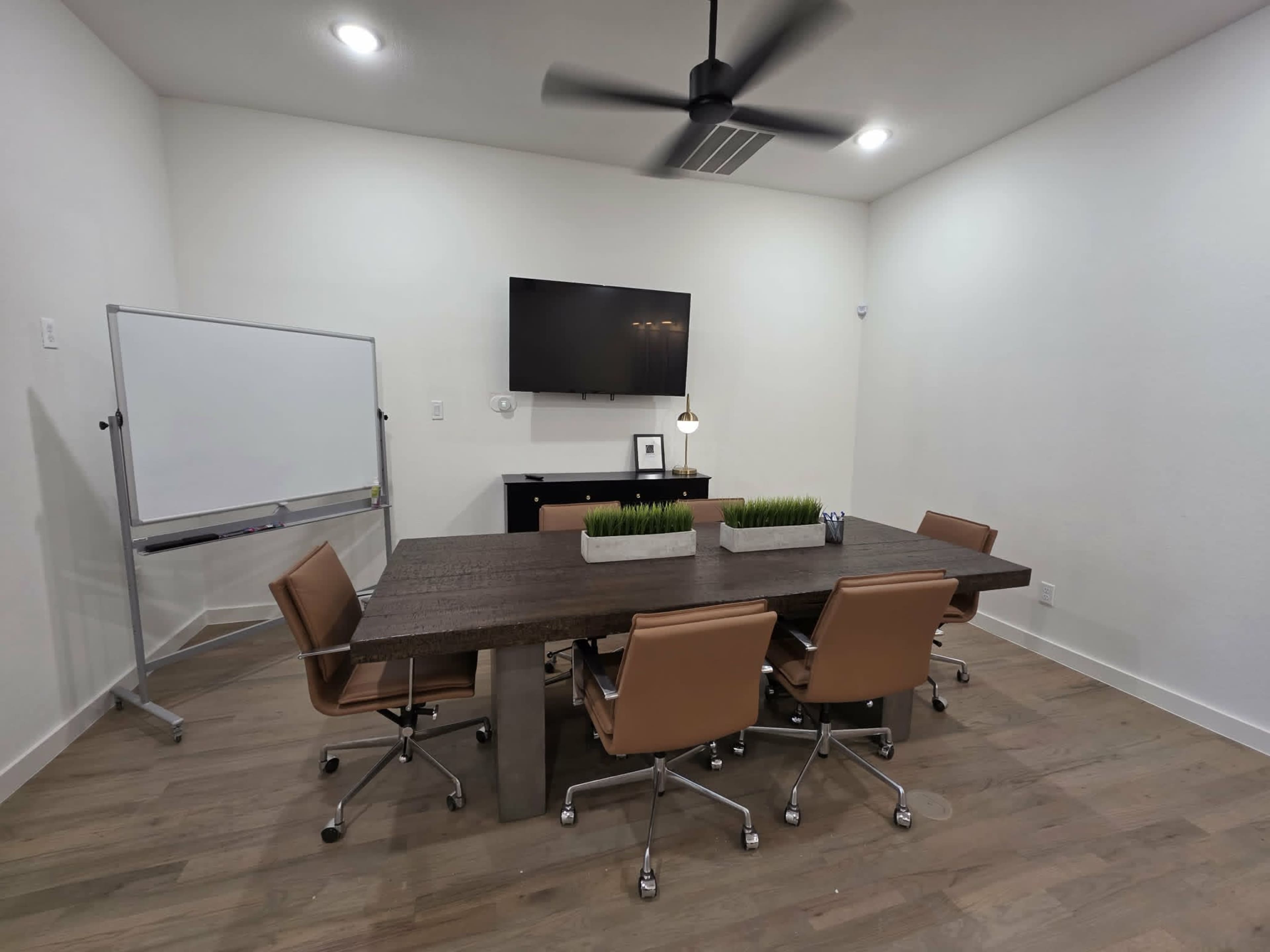 The image shows a modern conference room with a large wooden table, four brown office chairs, a wall-mounted television, a whiteboard, and a small planter on the table.