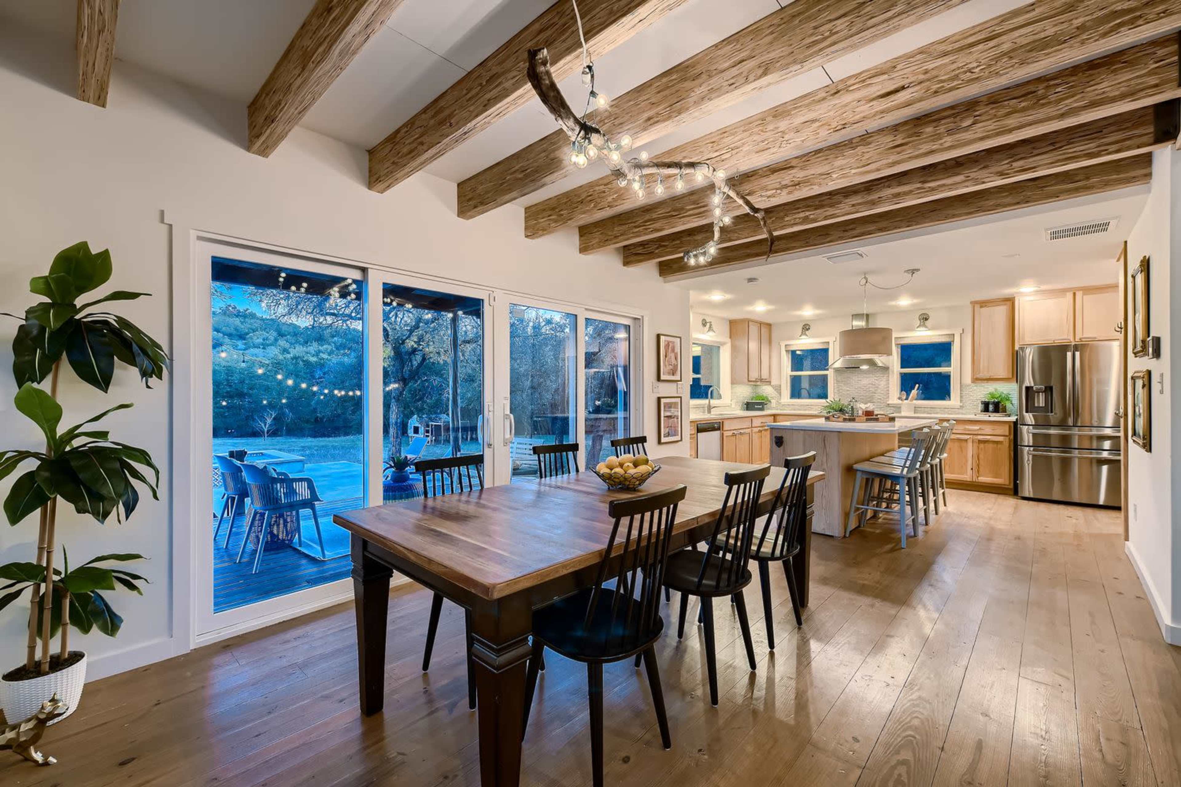 The image shows a spacious kitchen and dining area with wooden beams, a large table surrounded by black chairs, and large windows overlooking an outdoor patio.