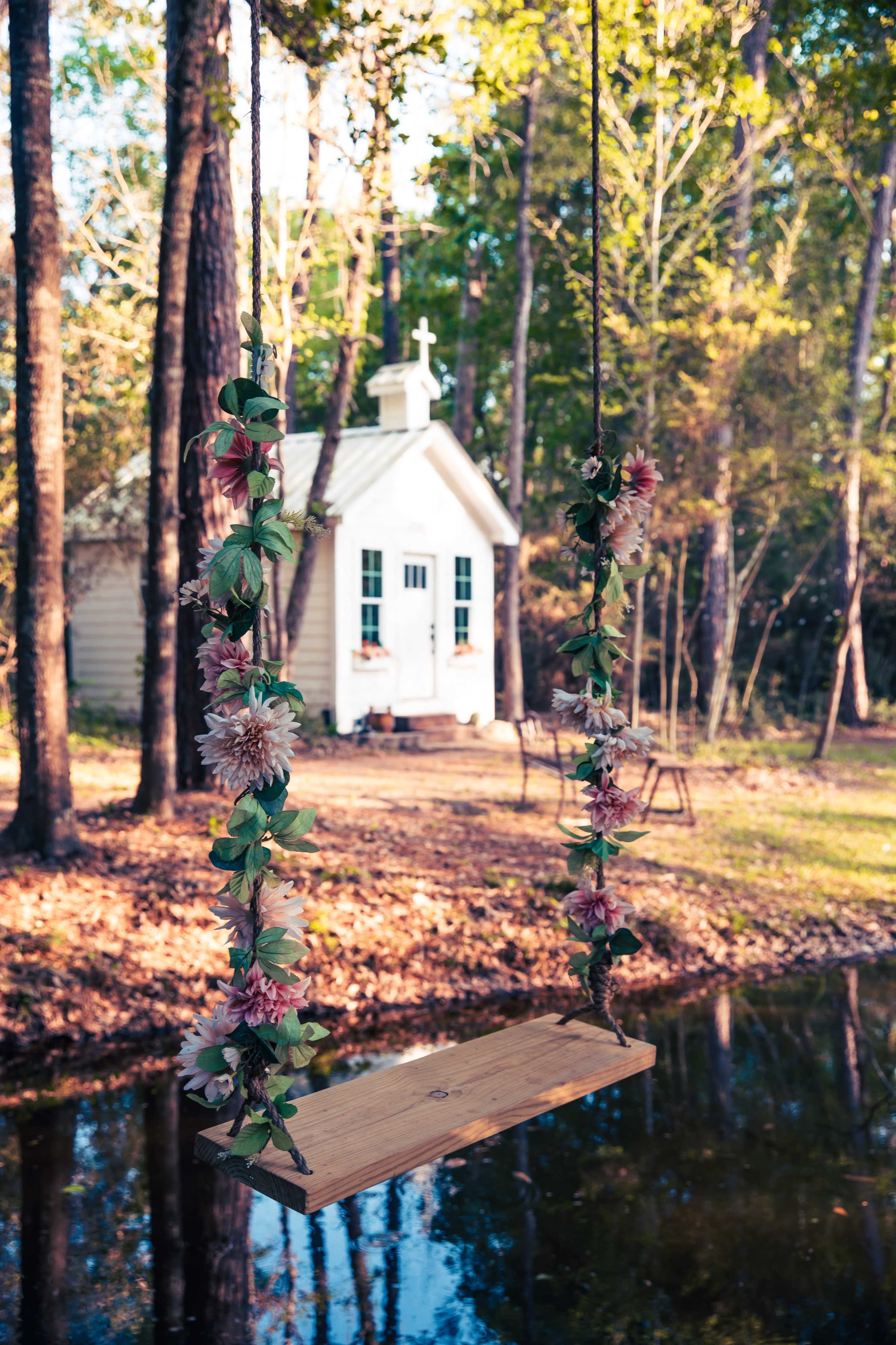 A wooden swing adorned with flowers hangs from a tree near a small, white chapel beside a calm pond.