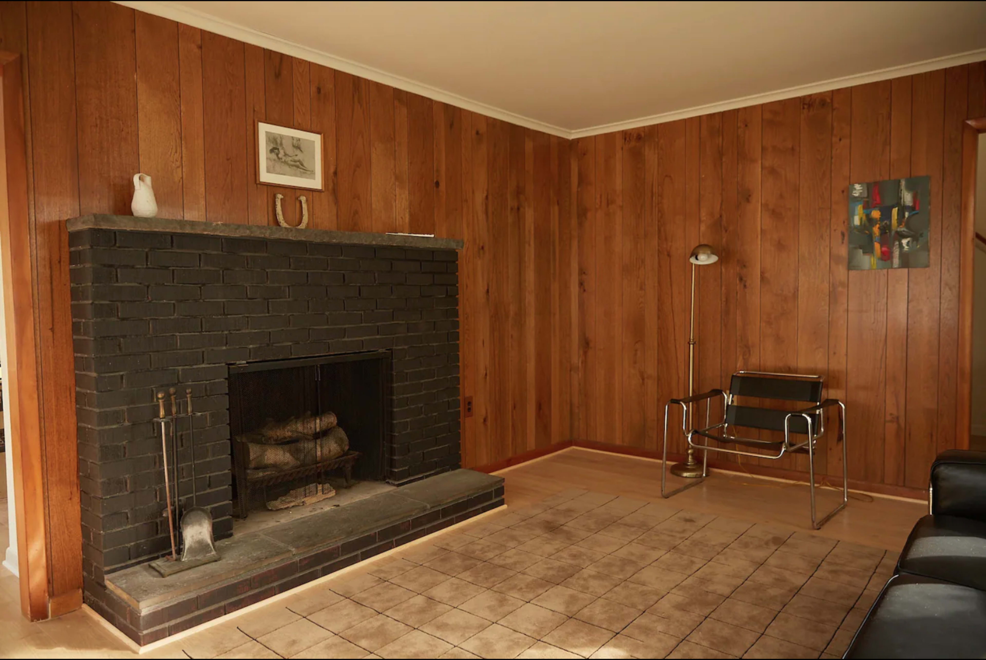 A living room with wooden paneled walls, a black brick fireplace, a floor lamp, a seating chair, and a tiled floor.
