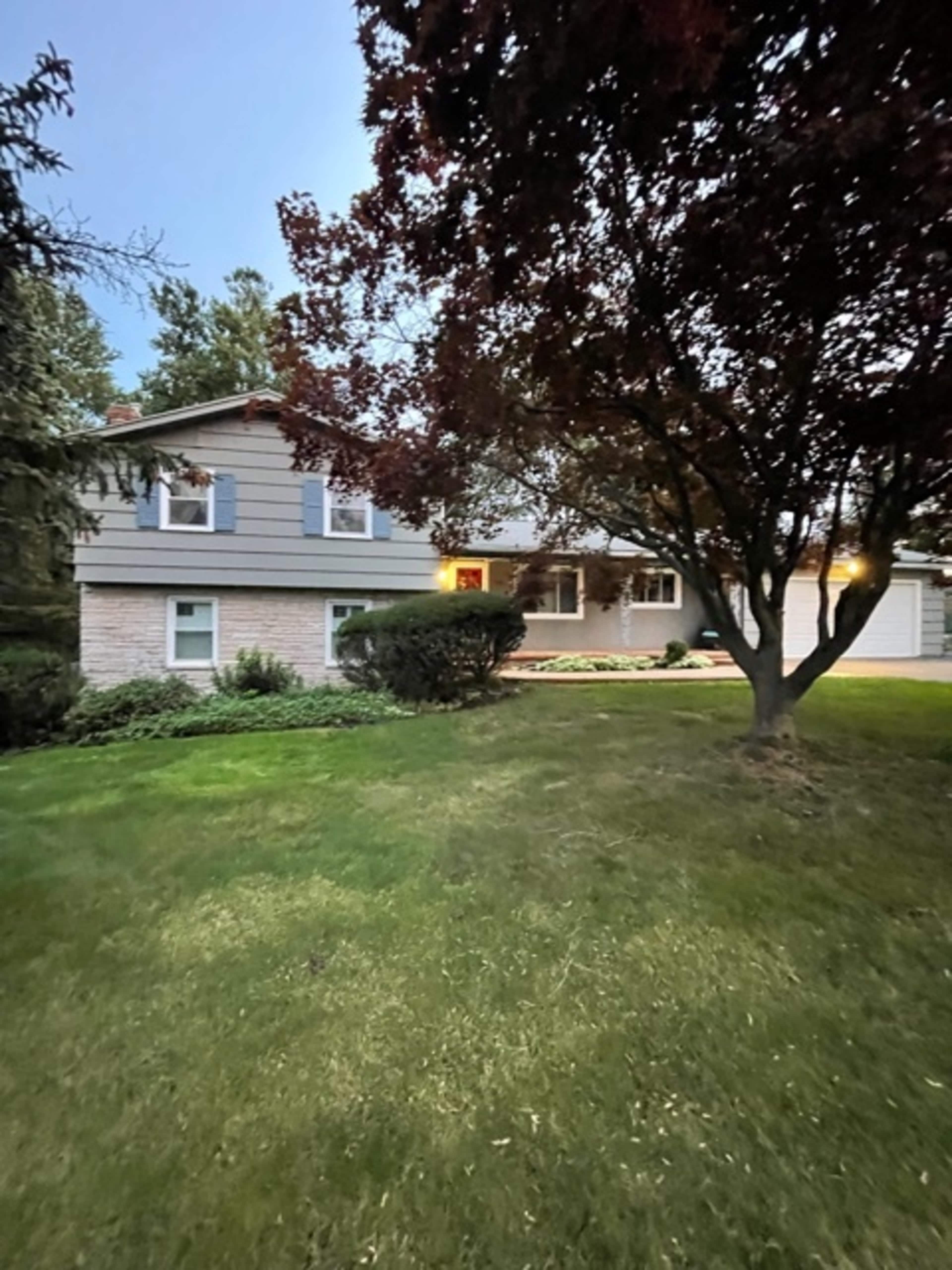 A two-story house with a manicured lawn and trees in the front yard is shown at dusk.