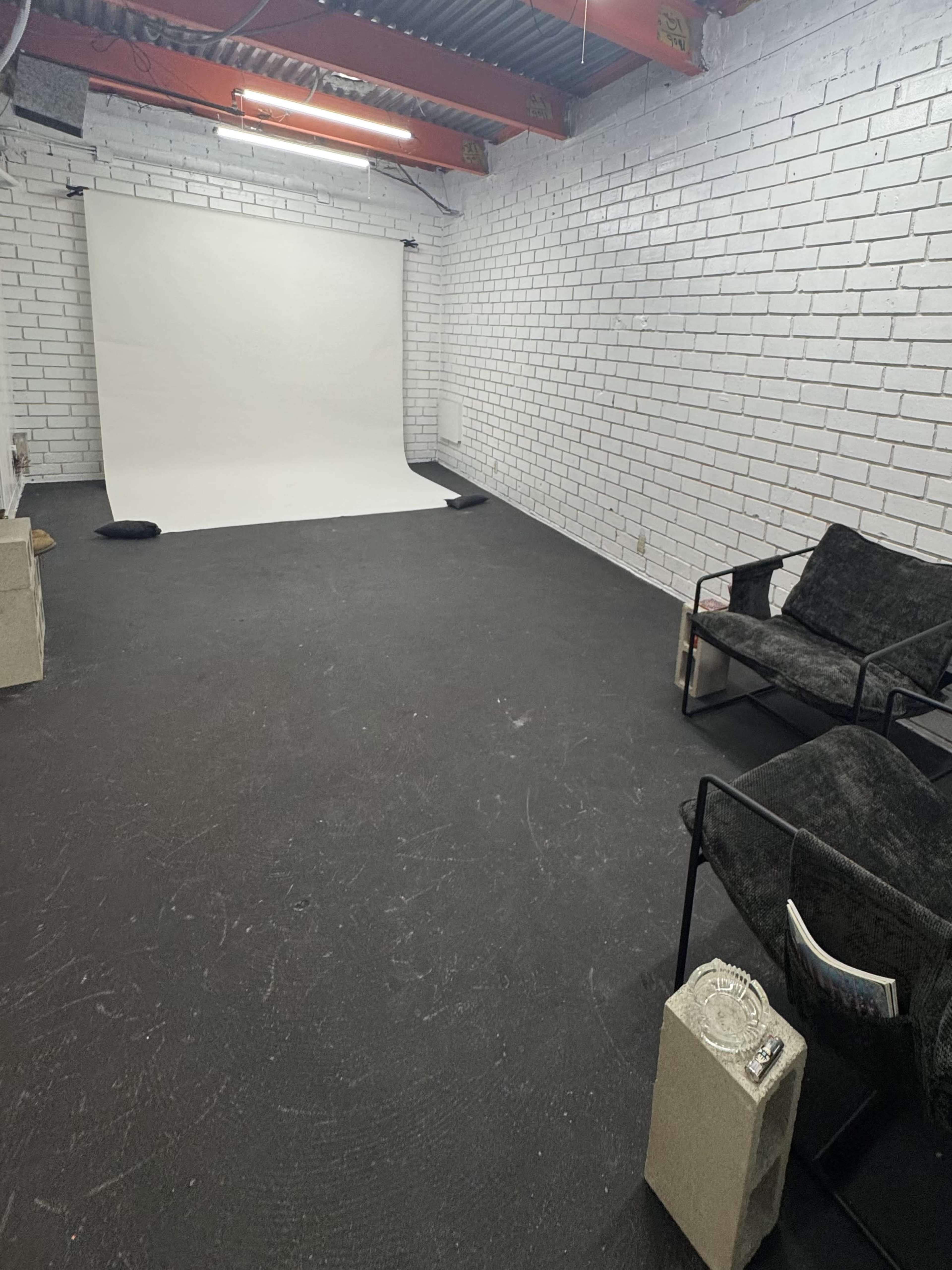 The image shows an empty room with white brick walls, a white backdrop on one side, a black floor, and two black chairs placed against a wall.