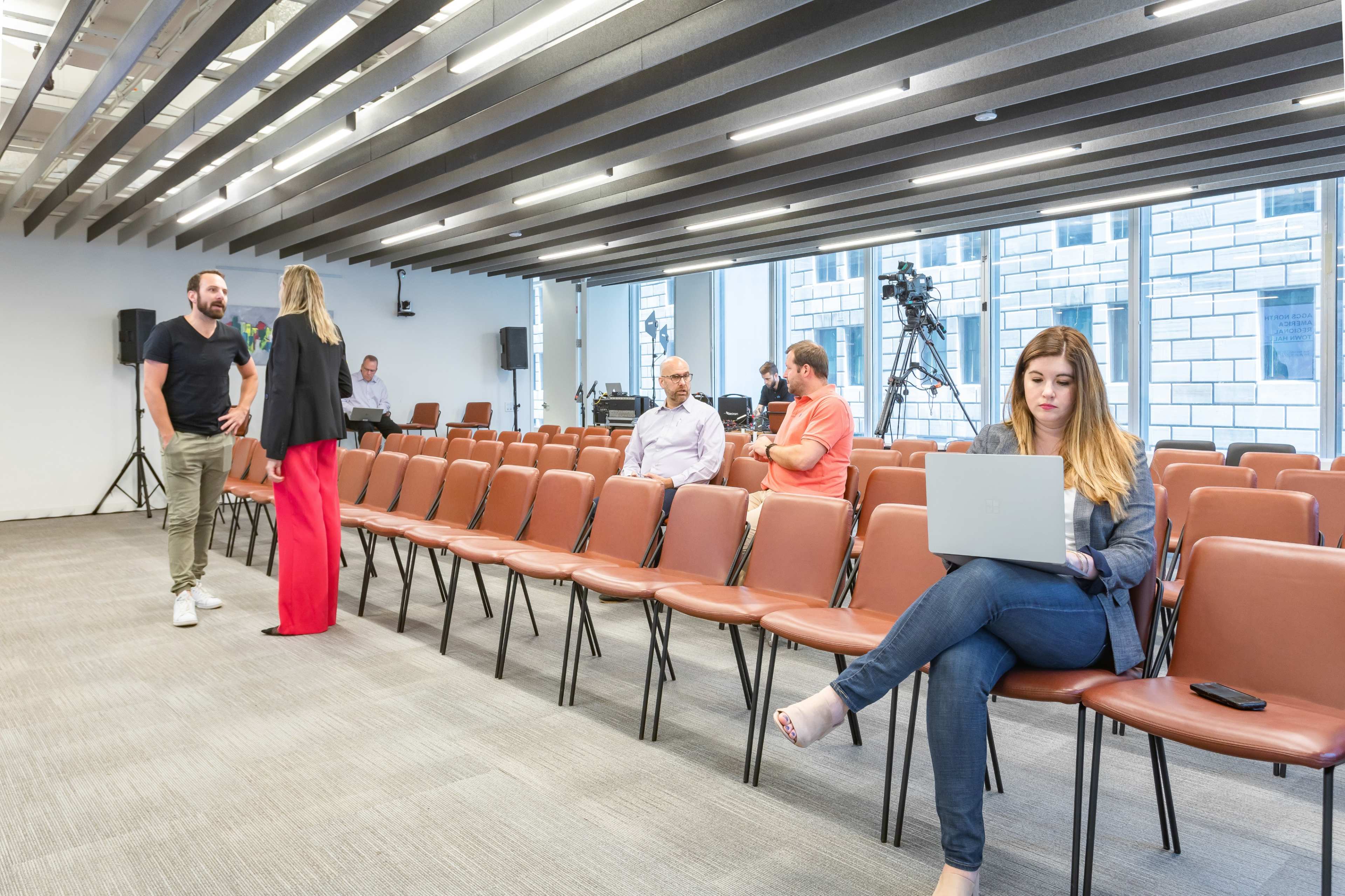 A largely empty conference room with rows of brown chairs, where a woman is sitting with a laptop and a few people are conversing nearby.
