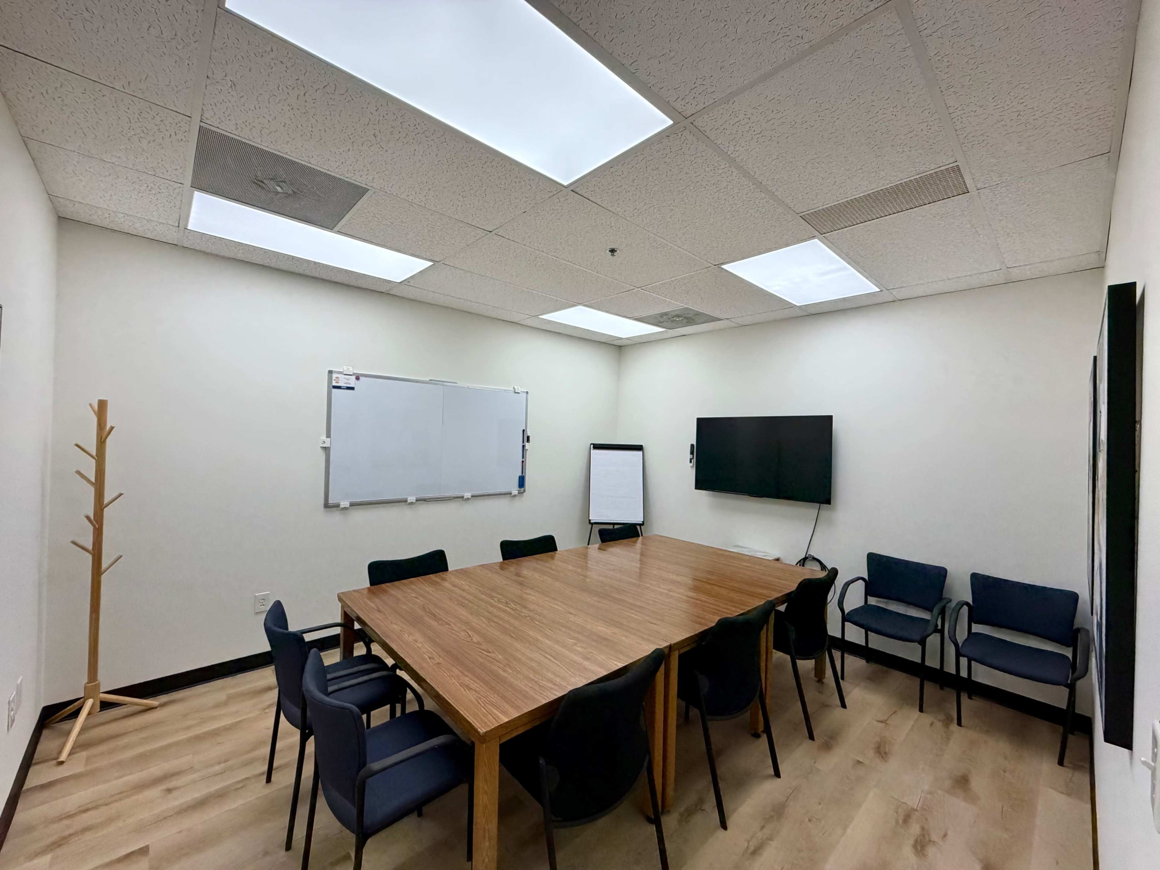 A conference room features a large wooden table surrounded by eight chairs, a whiteboard, and a television mounted on the wall.