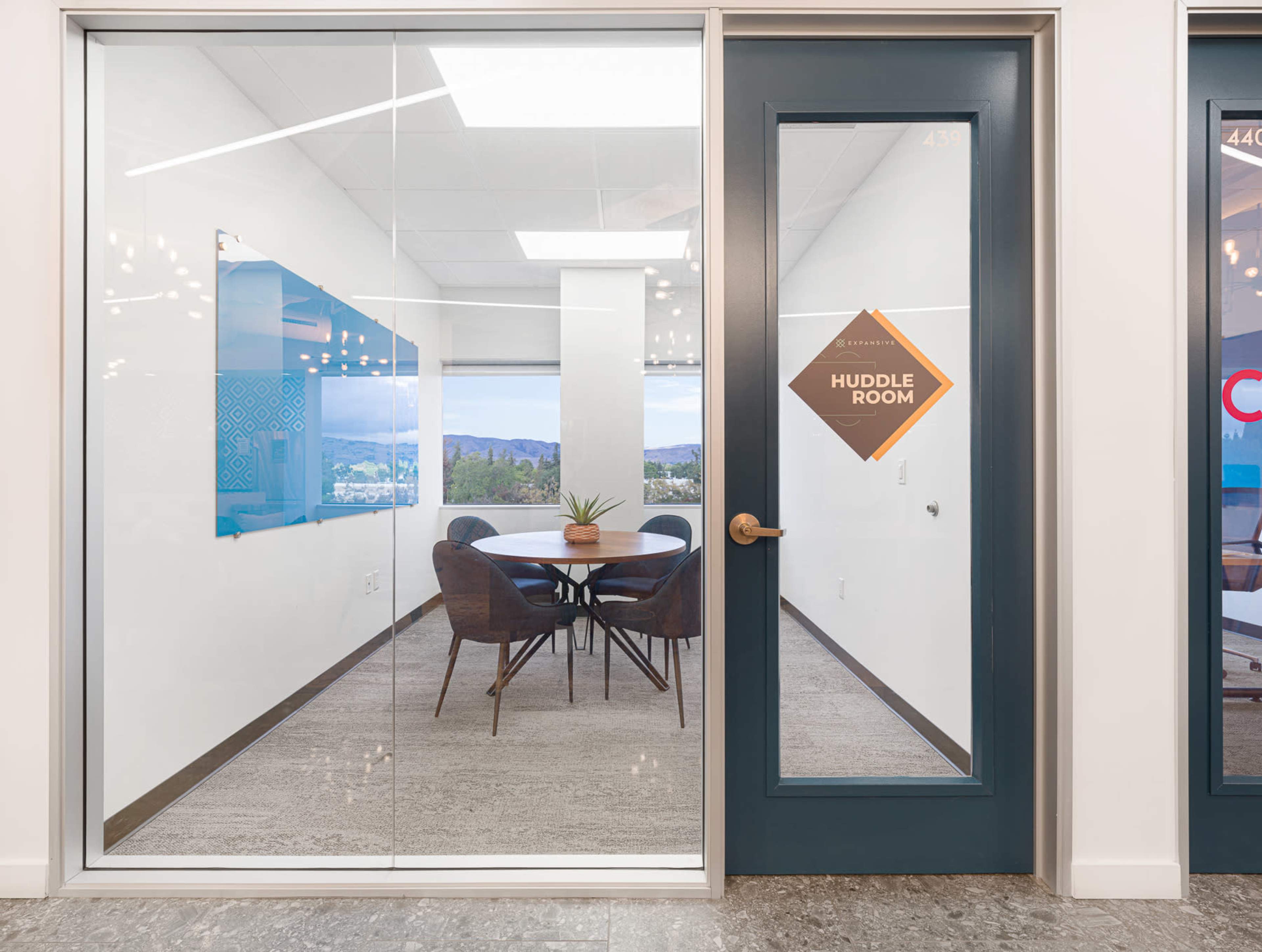 The image shows a glass-walled huddle room with a round table and chairs, featuring a view of hills in the background.