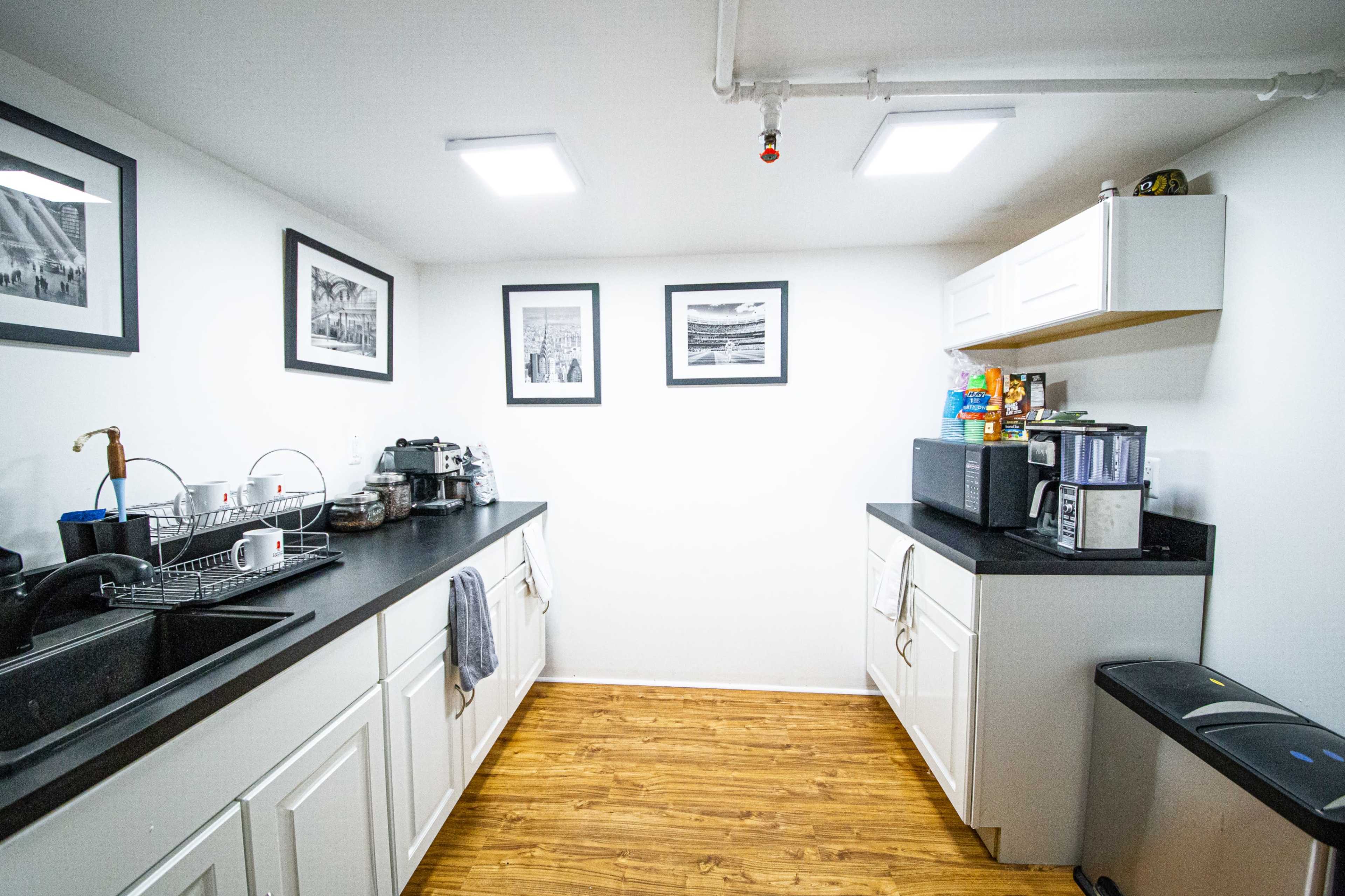 The image shows a compact kitchen with white cabinets, black countertops, and several framed pictures on the walls.