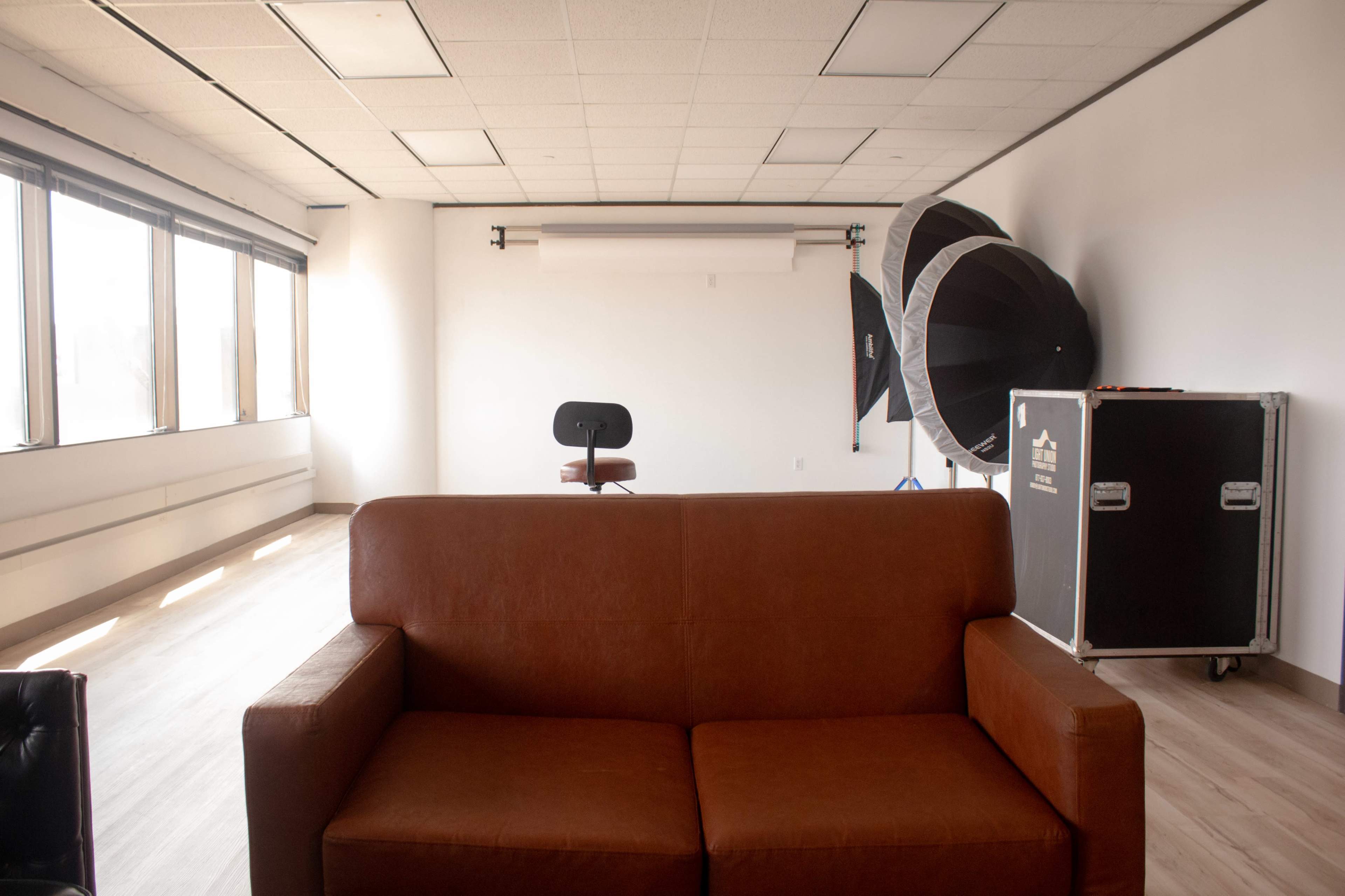 The image shows an empty room with a brown couch facing a wooden chair and a backdrop setup, alongside two large softboxes.