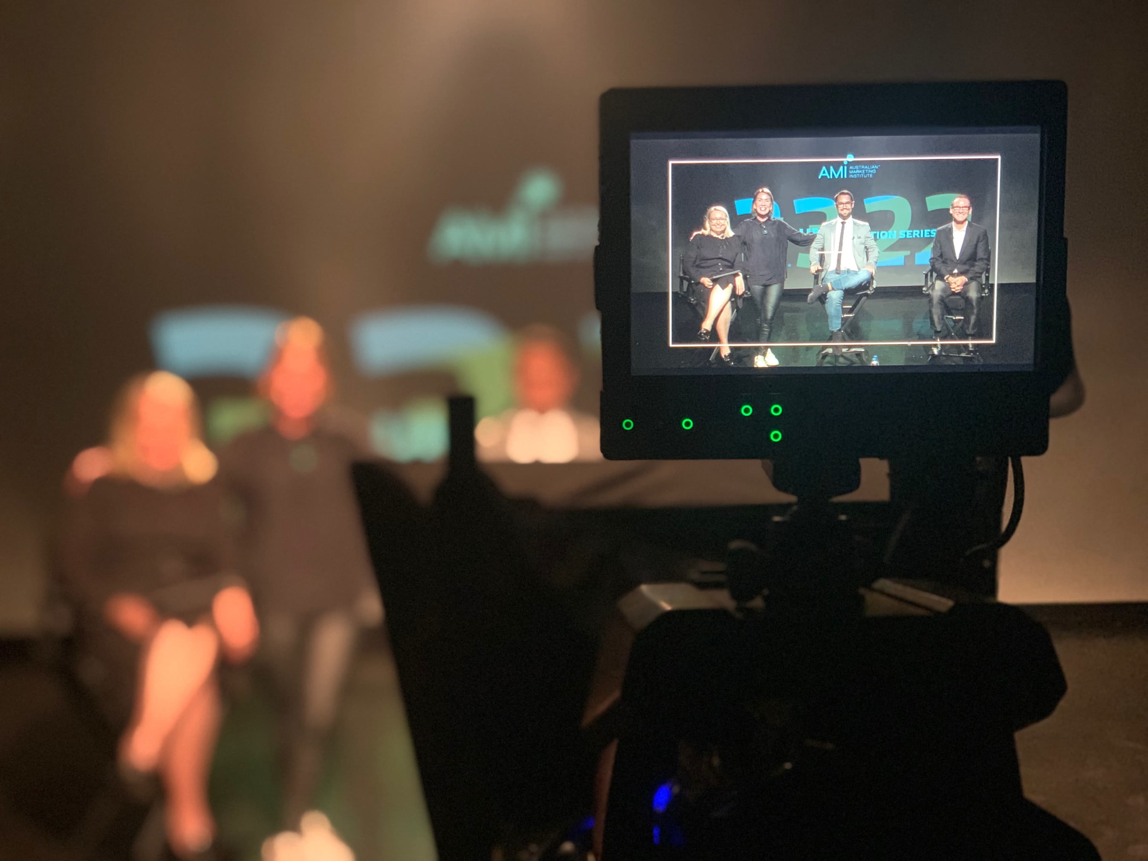 A camera captures a group of four people seated on a stage during a presentation, with a screen displaying branded graphics in the background.
