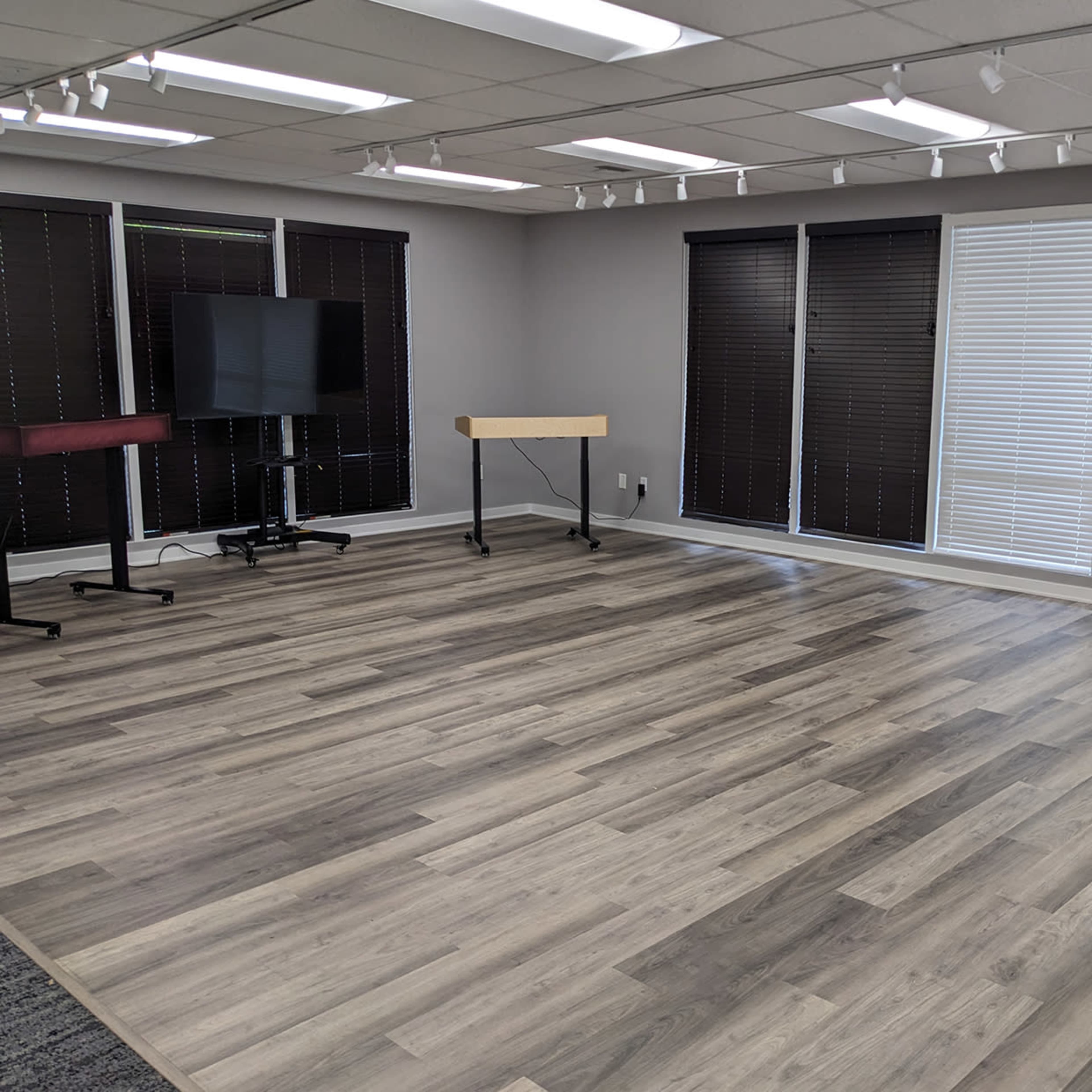 A spacious room with wooden flooring, equipped with a television on a stand, a small table on wheels, and several window blinds.