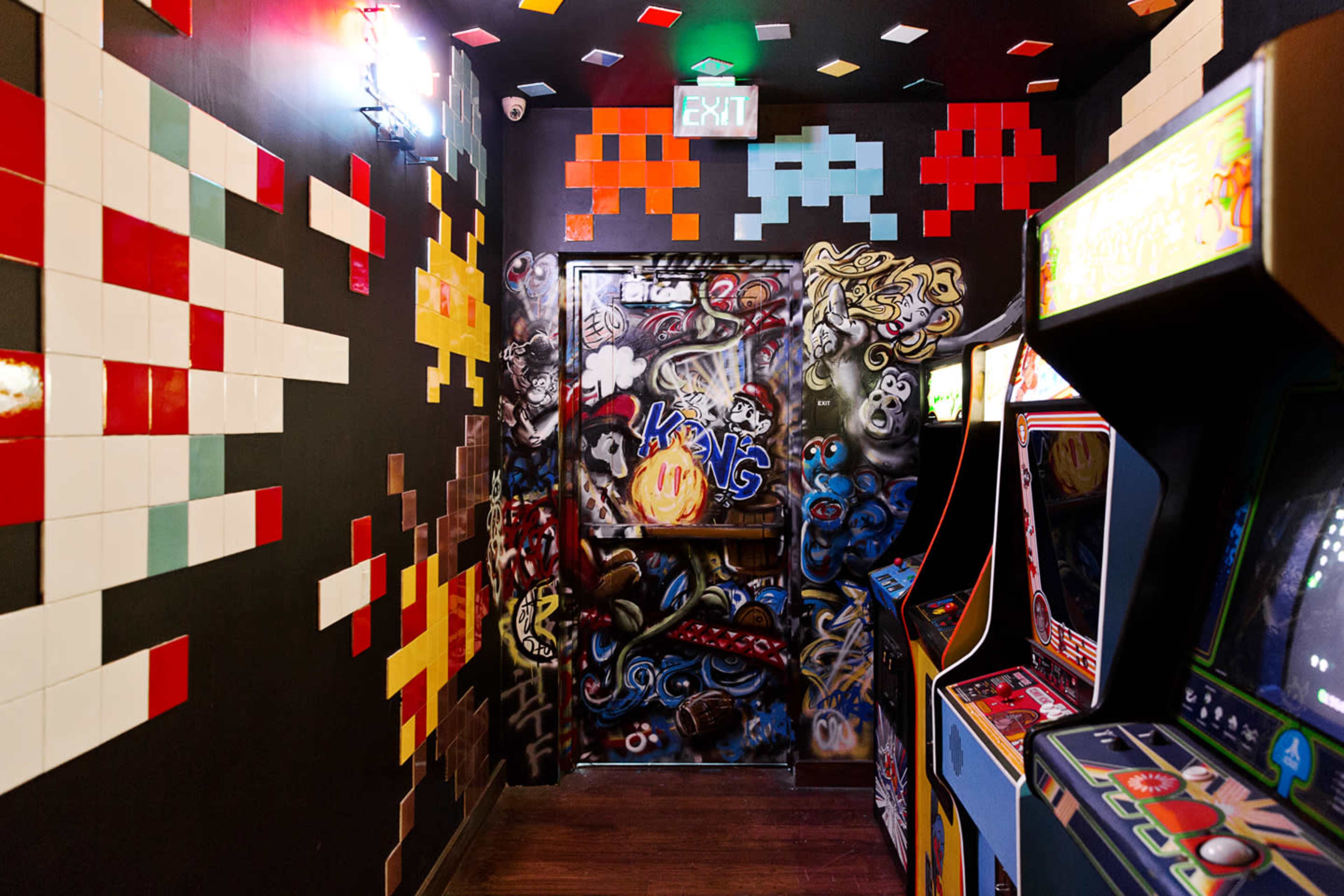 The image shows a vibrant arcade hallway decorated with pixel art and graffiti, featuring vintage arcade machines lined up against the walls.