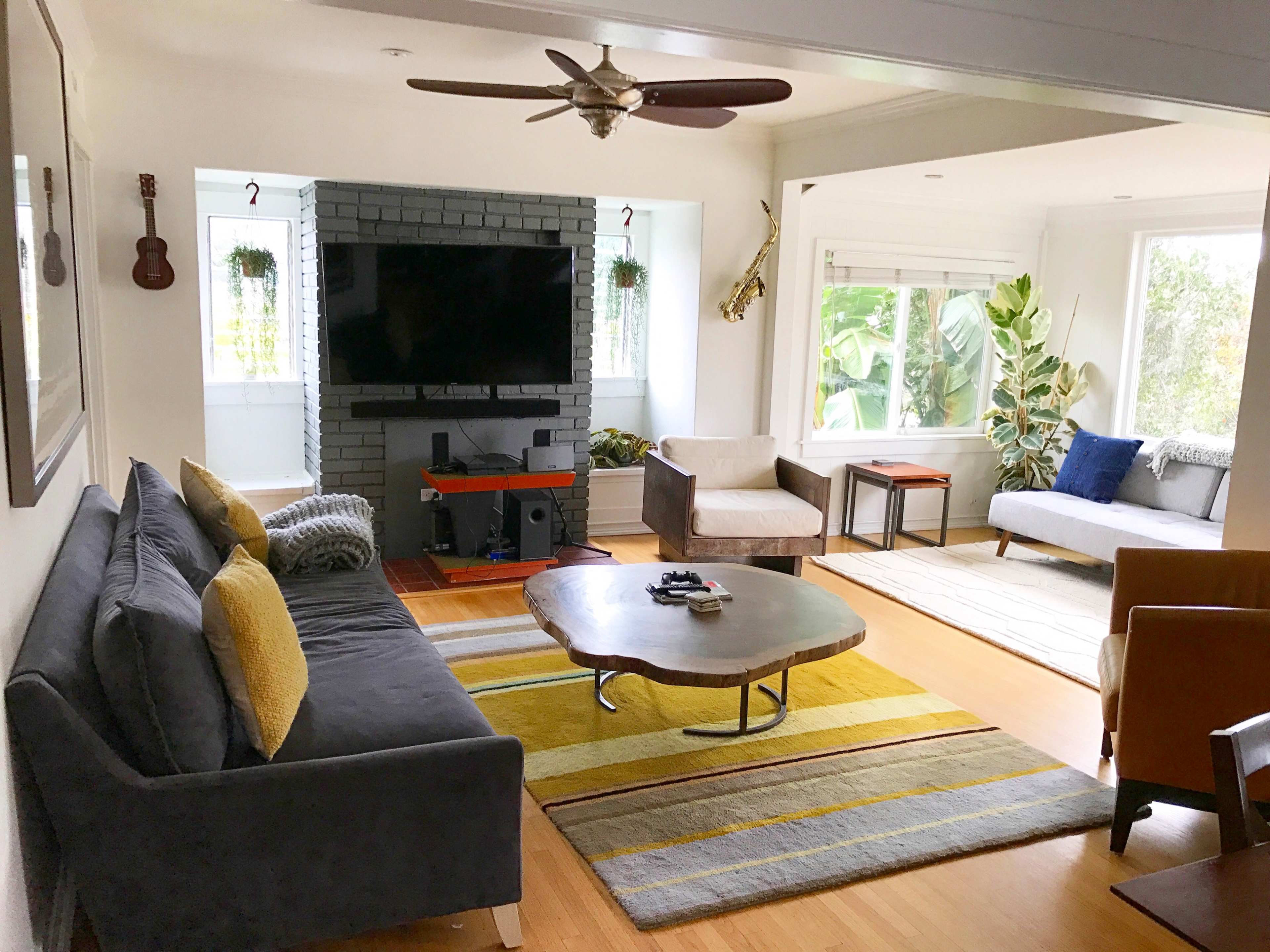 The image shows a modern living room featuring a gray sofa, two armchairs, a coffee table, a television on a black-brick wall, and windows with plants.