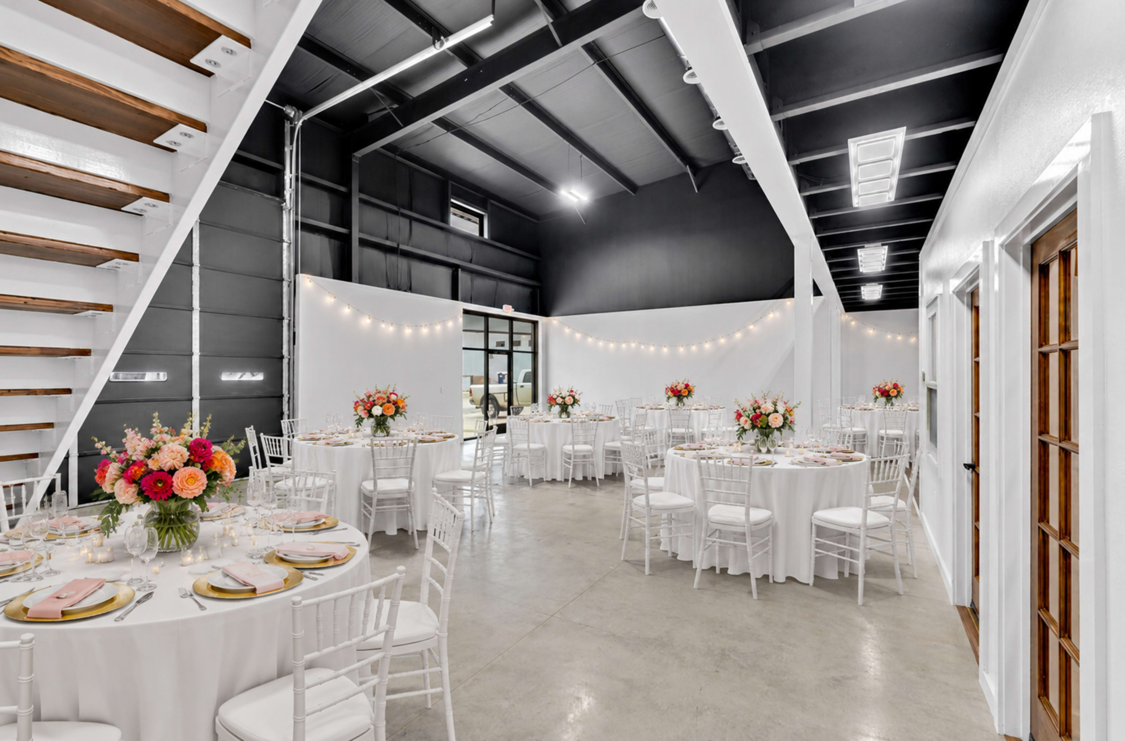The interior of a spaciously decorated event venue features round tables set with floral centerpieces and white chairs, illuminated by soft string lights.
