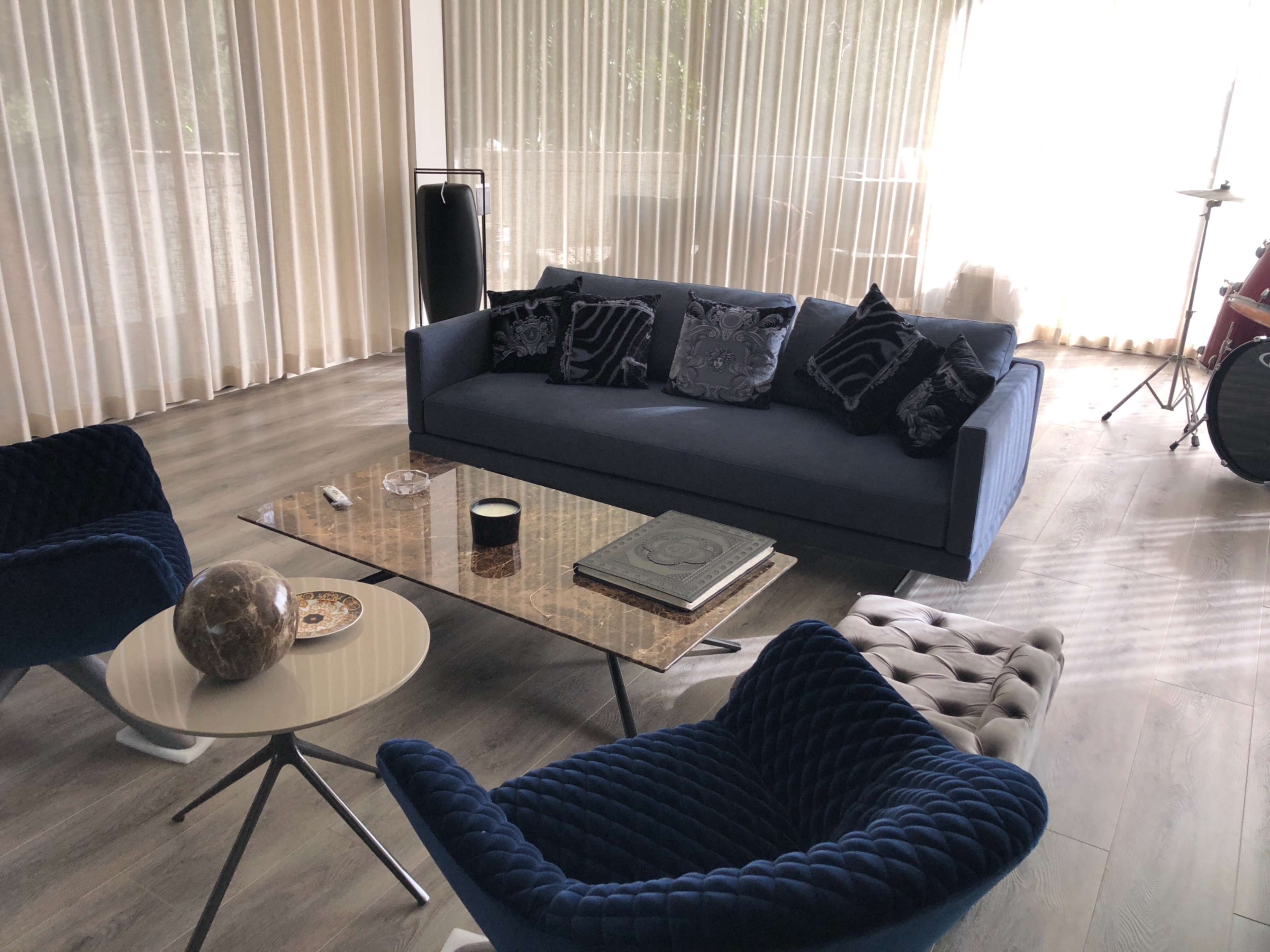The image shows a modern living room with a blue sofa, patterned cushions, a glass coffee table, and two blue armchairs, featuring large windows covered by sheer curtains.