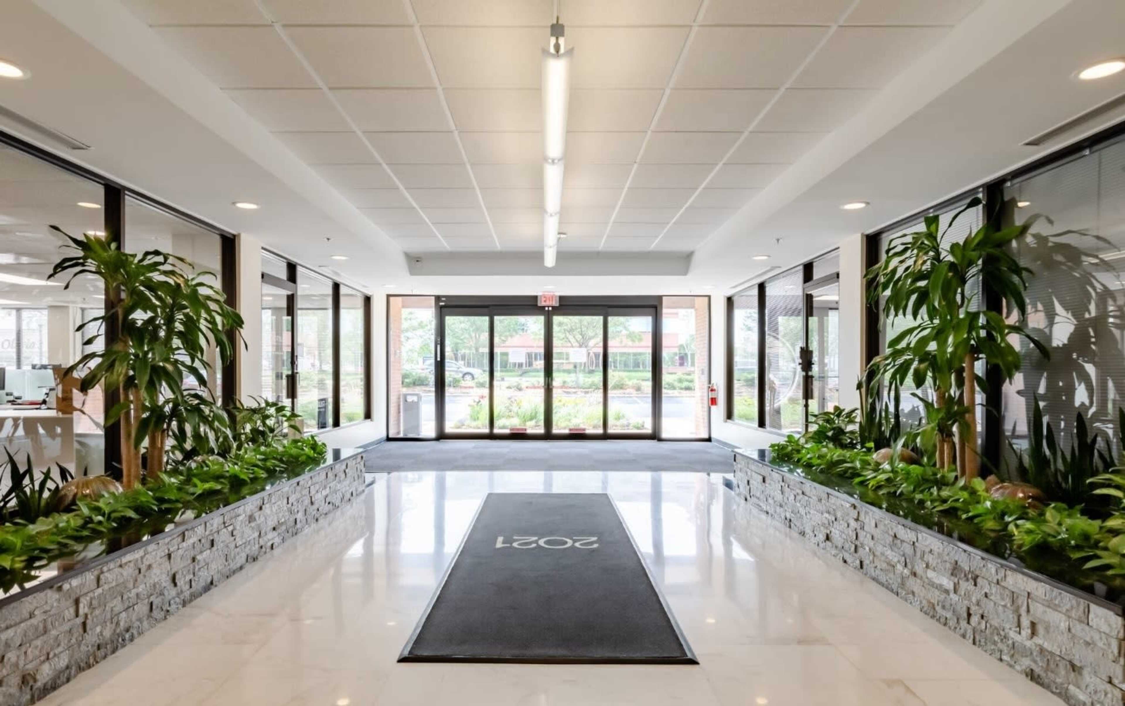 A spacious hallway with large windows, a stone-lined planter on each side, and a carpet mat displaying the number "2021" at the entrance.