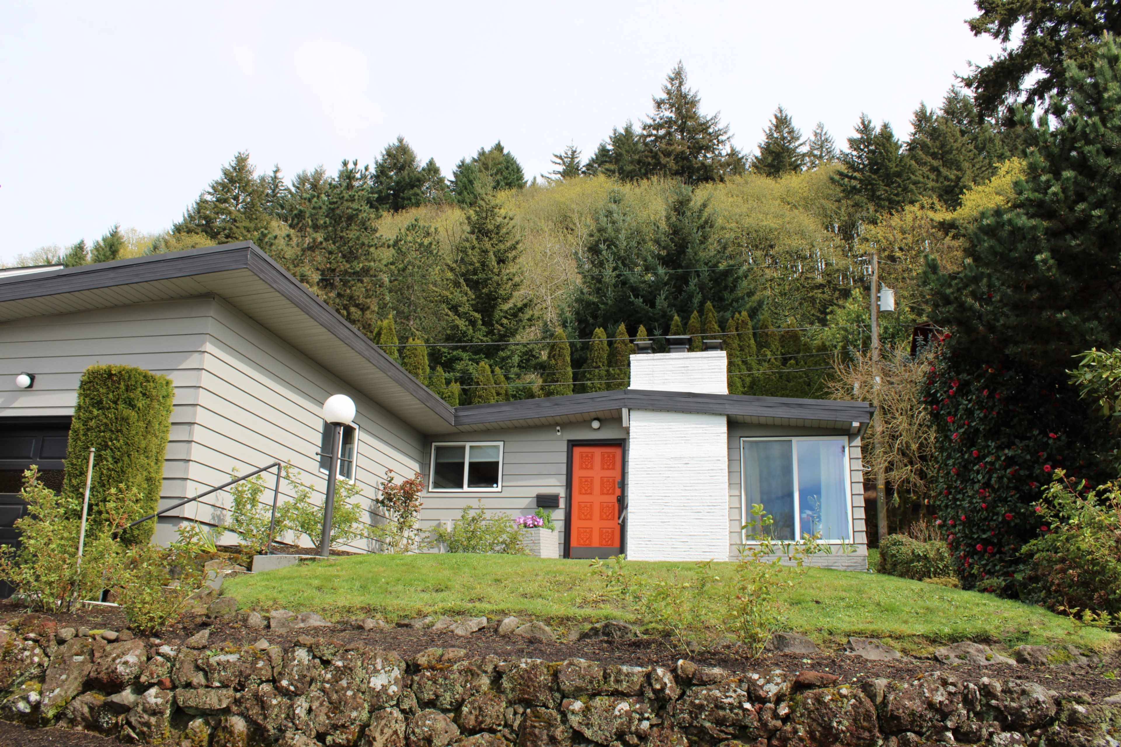 A contemporary house with a bright orange front door sits on a grassy lawn surrounded by trees and shrubs.