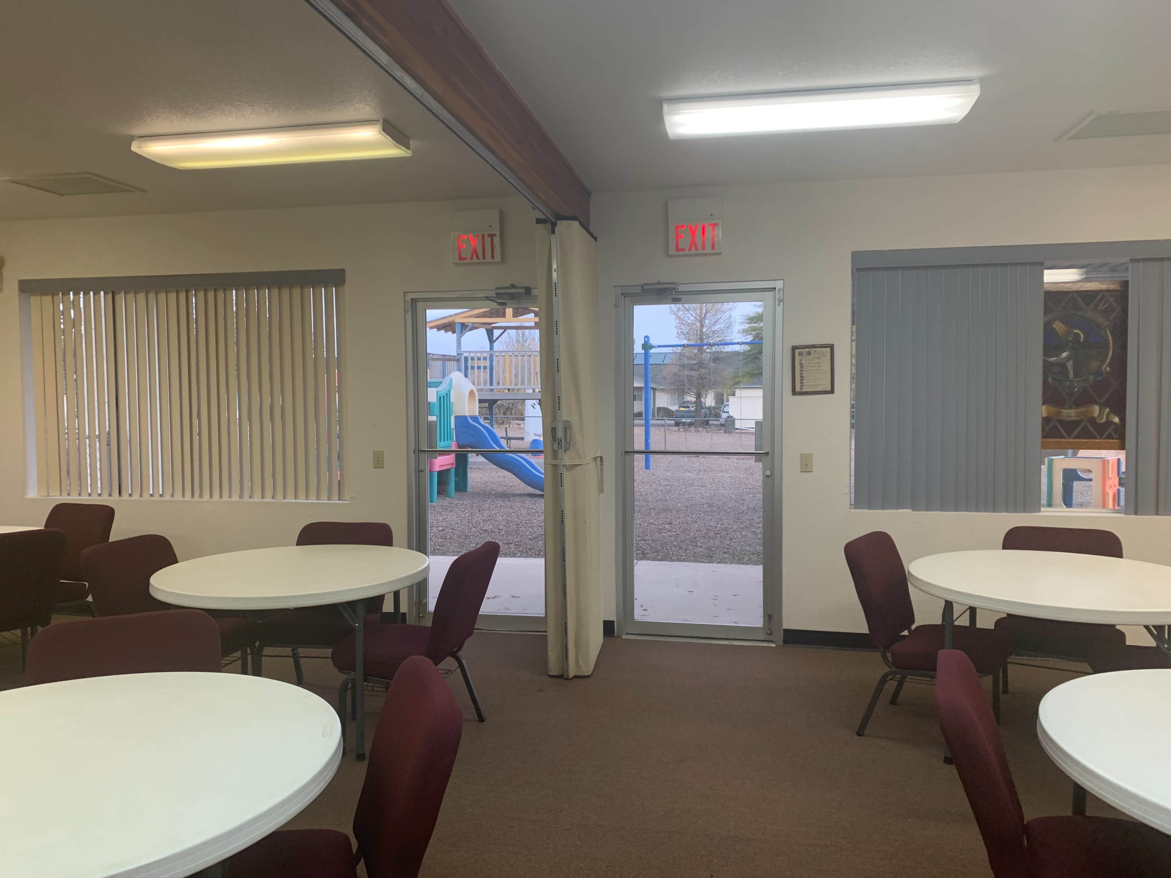A community room with round tables and chairs, leading to two exit doors that open to a playground area outside.