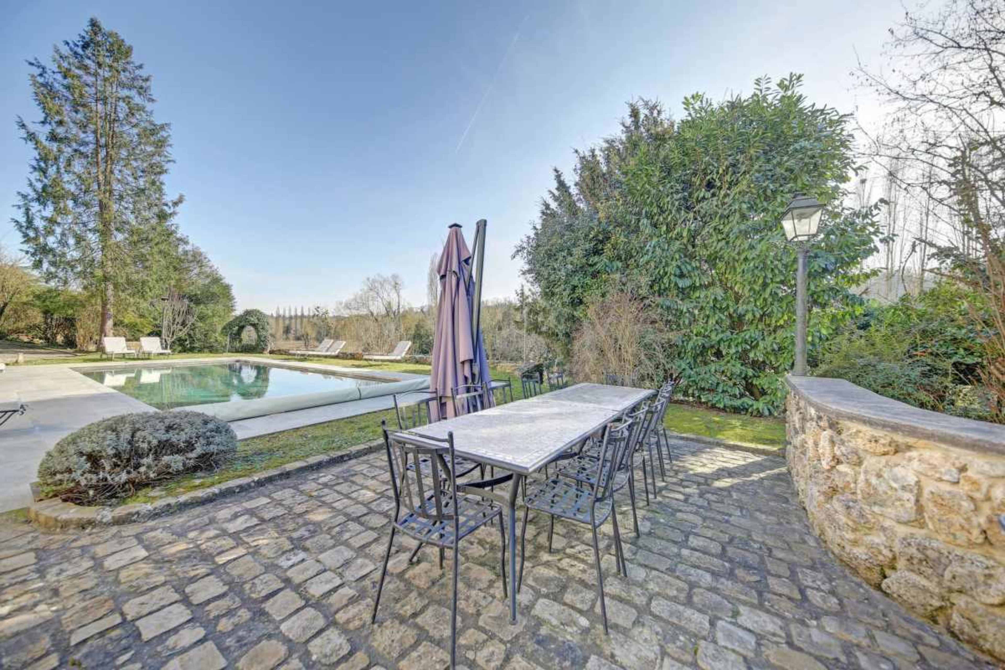 The image shows an outdoor dining area with a large table and chairs near a swimming pool surrounded by greenery.