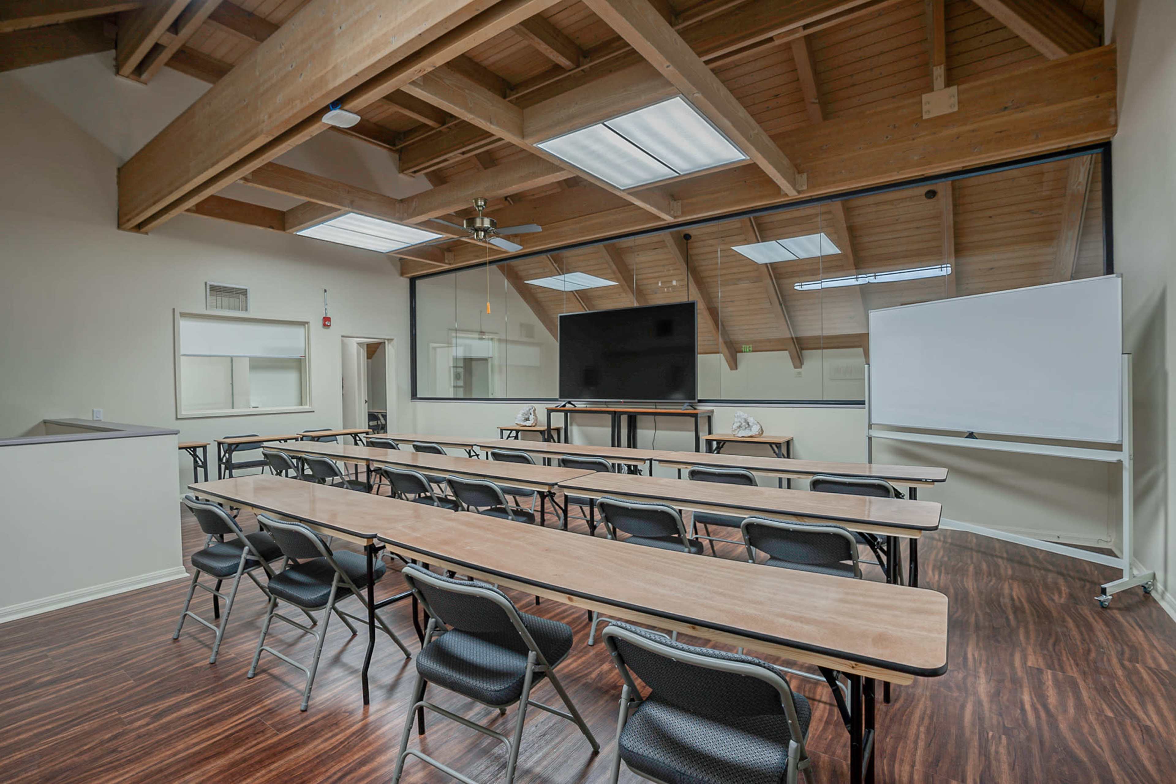A classroom features several rows of tables and chairs, with a large screen and whiteboard at the front, under a wooden beam ceiling.